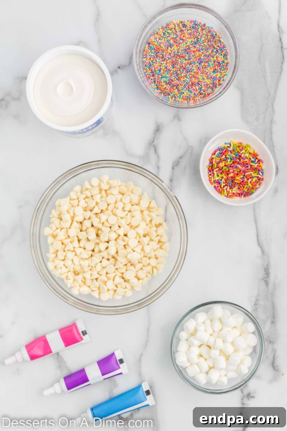Enchanted Unicorn Swirl Fudge 3 A flat lay photograph showcasing the five key ingredients for Unicorn Fudge: white chocolate chips, a container of white frosting, various food colorings, mini marshmallows, and rainbow sprinkles.