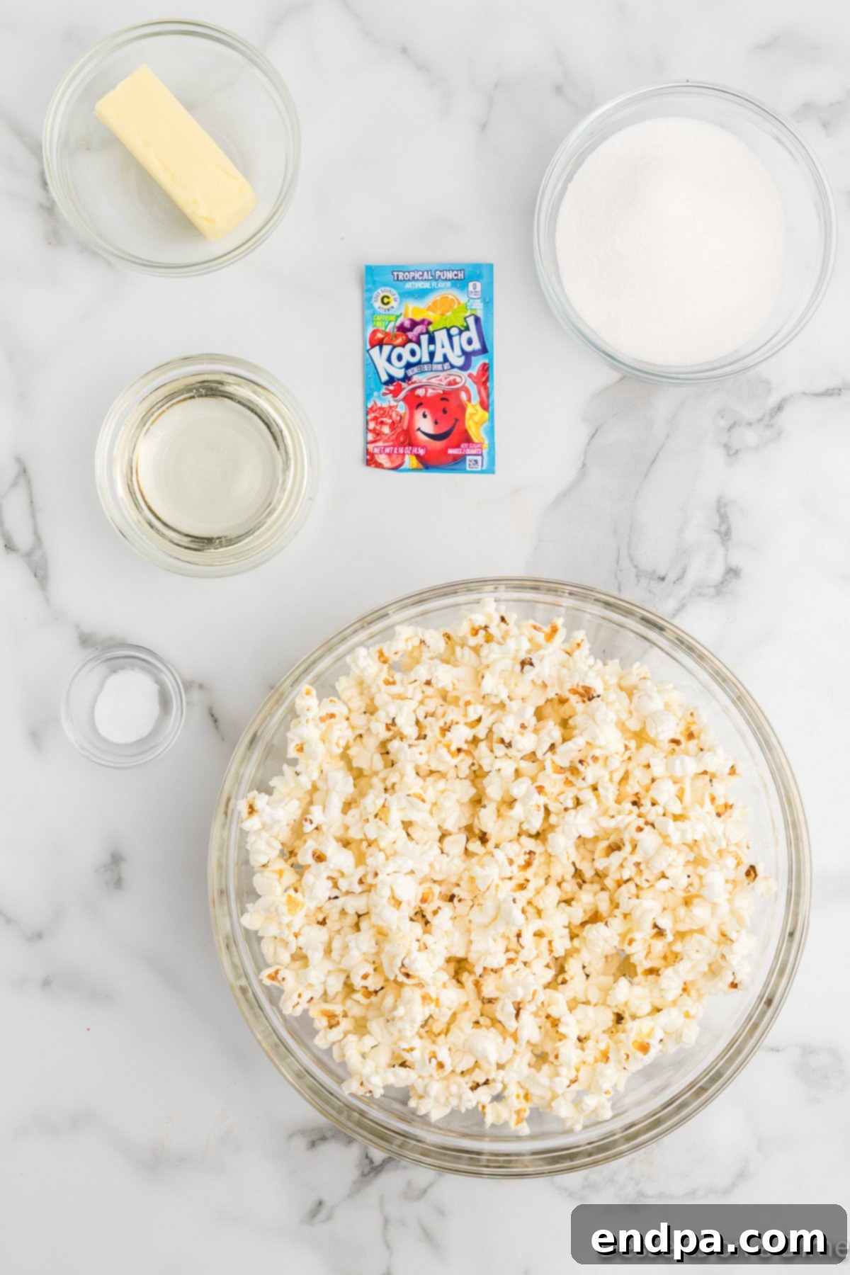 A flat lay of ingredients for Kool-Aid popcorn, including popped popcorn, granulated sugar, light corn syrup, butter, a Kool-Aid packet, and baking soda.