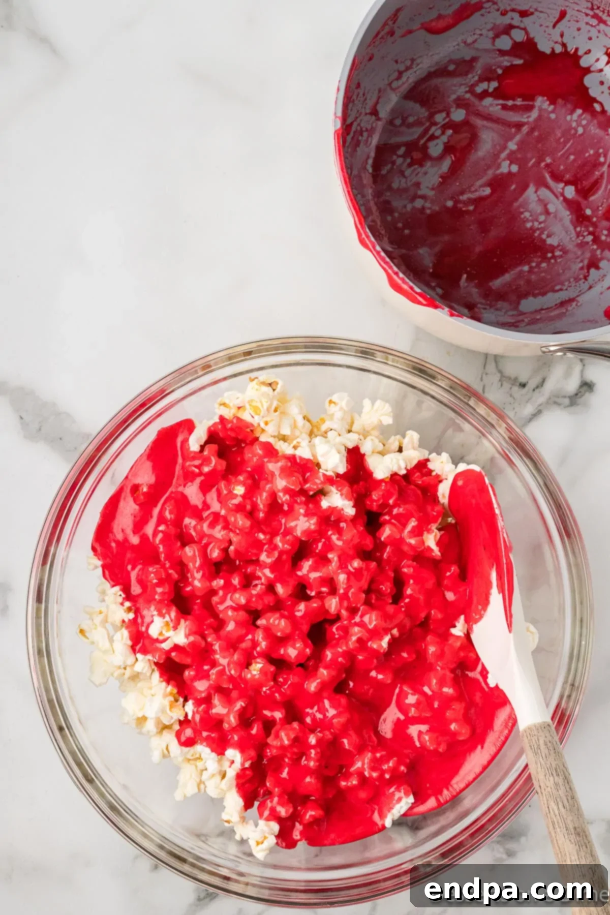 The colorful Kool-Aid mixture being poured carefully over the popped popcorn in a large bowl.