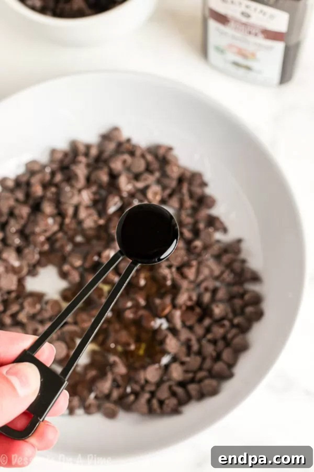 The Ultimate Homemade Ice Cream Cake 3 A teaspoon pouring vanilla extract into a bowl containing chocolate chips and corn syrup, preparing the hot fudge sauce.