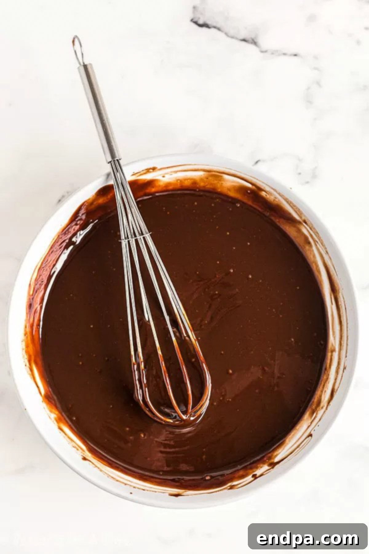 The Ultimate Homemade Ice Cream Cake 4 A whisk stirring a bowl of freshly melted chocolate fudge, creating a smooth and glossy texture.