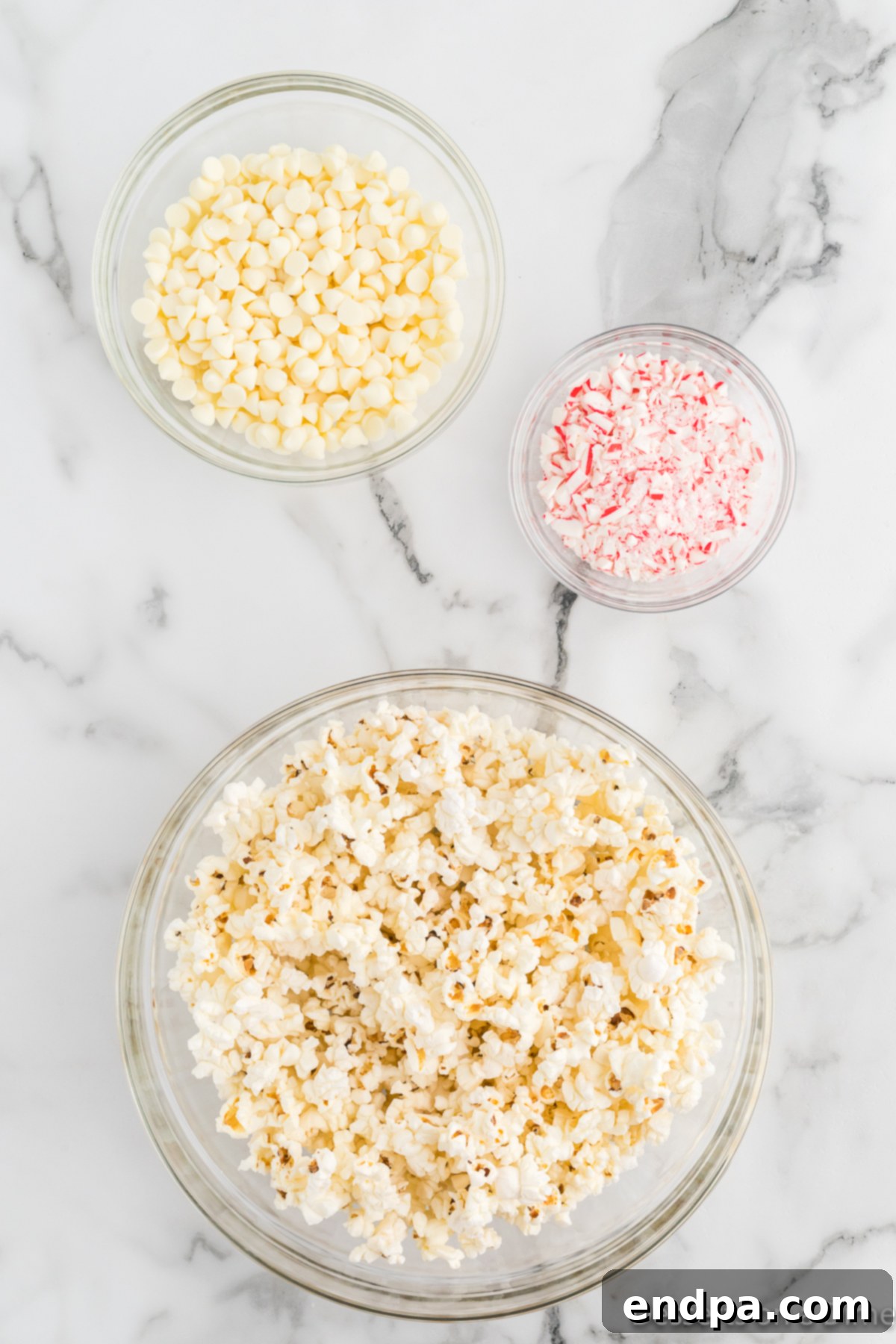 Peppermint Pop Crunch 3 Essential ingredients for Candy Cane Popcorn, including popped popcorn, white chocolate chips, and peppermint candy canes.