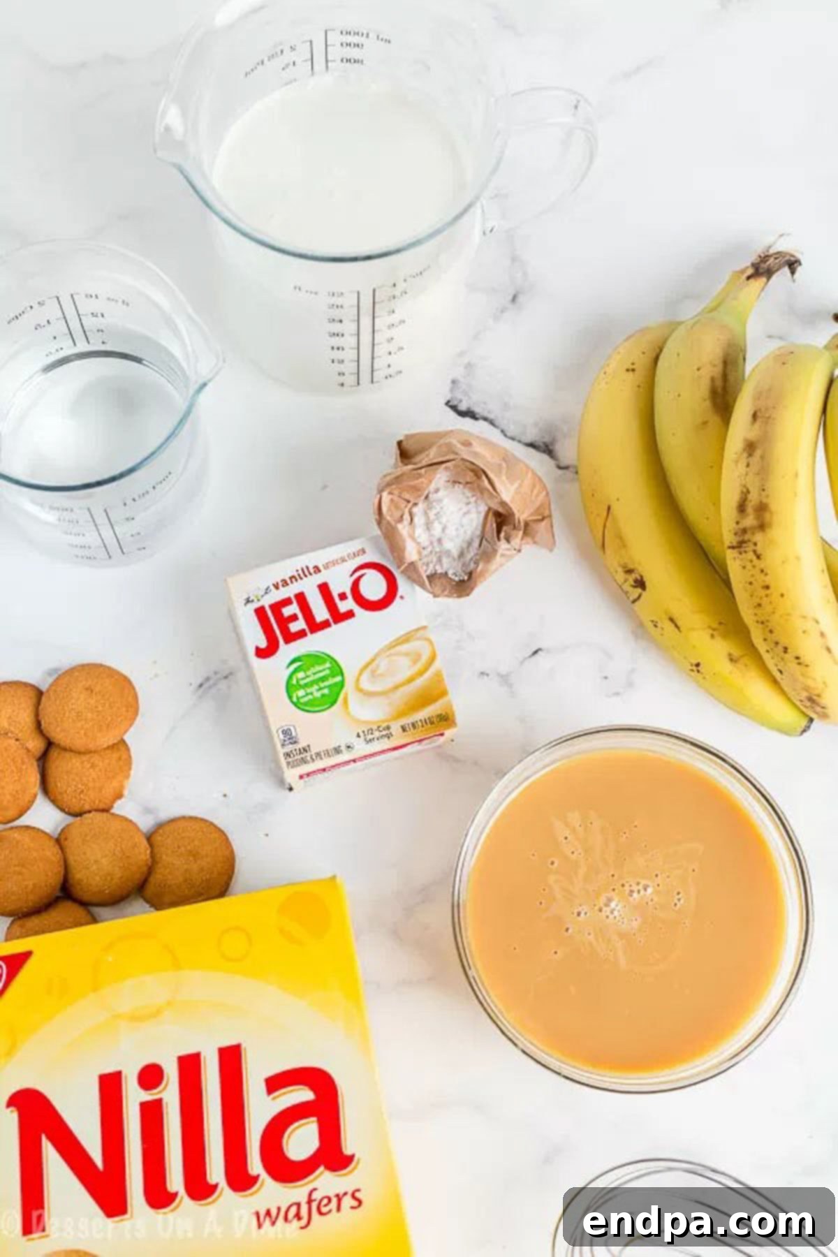 A flat lay image showing all the essential ingredients for banana pudding: sweetened condensed milk, instant vanilla pudding mix, heavy cream, fresh bananas, and Nilla wafers.