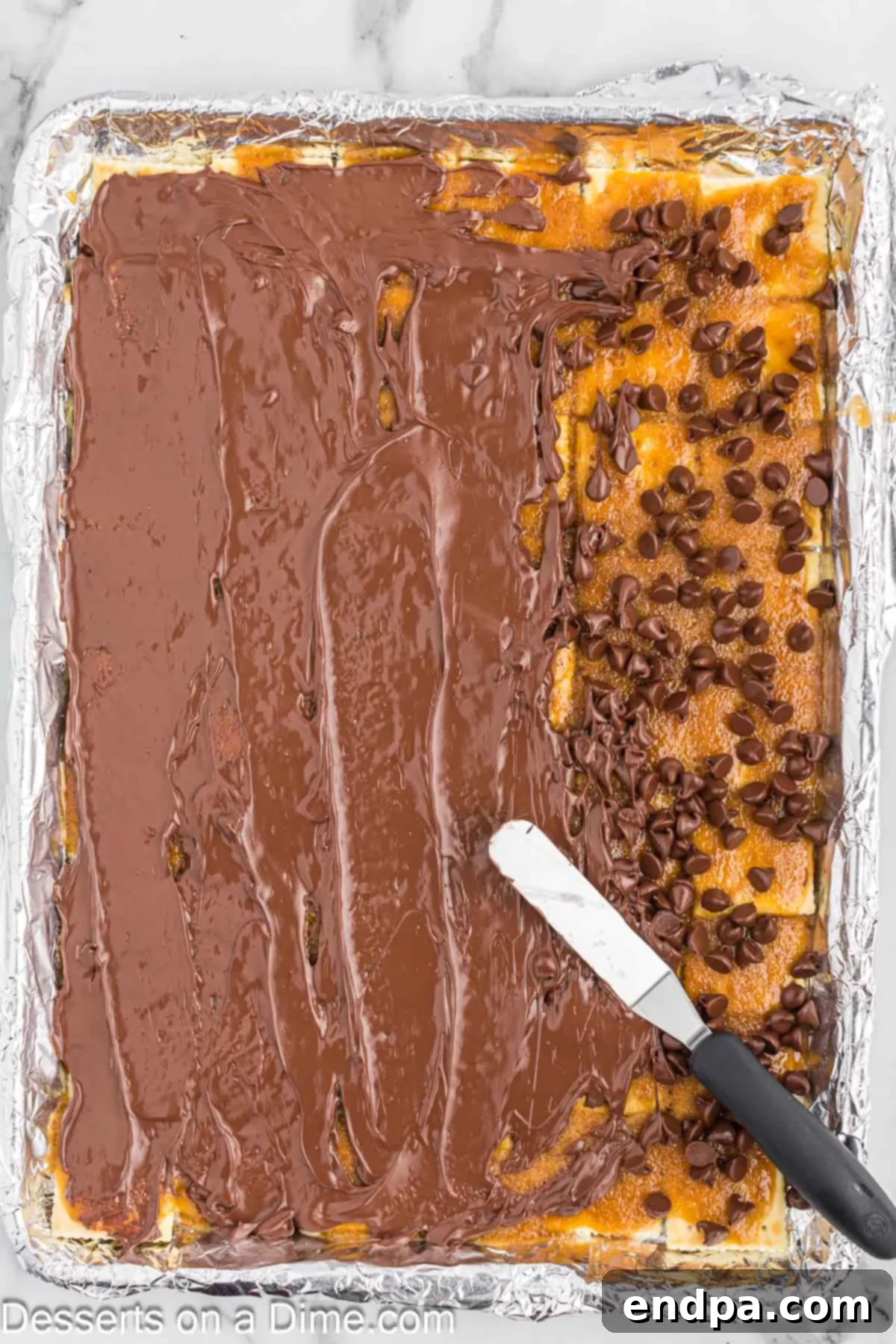A spatula smoothly spreading the melted chocolate chips over the warm toffee and cracker base, creating an even, glossy layer for the Christmas Crack.