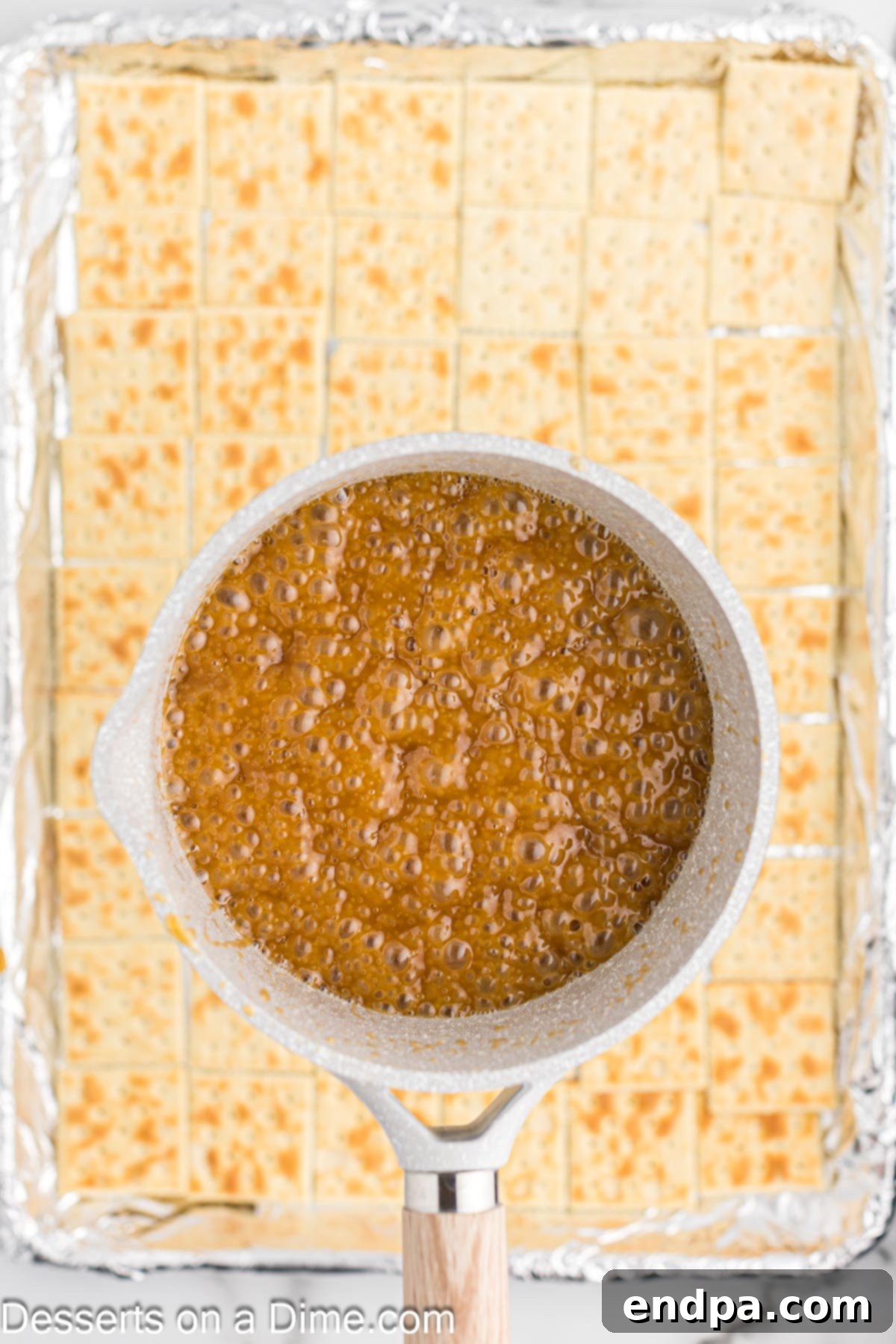Hot, bubbly toffee mixture being carefully poured from a saucepan over the arranged saltine crackers on a baking sheet, ensuring full coverage.