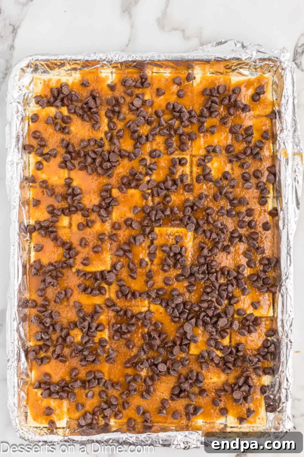A generous layer of semi-sweet chocolate chips sprinkled evenly over the hot, freshly baked toffee-coated crackers on a baking sheet.