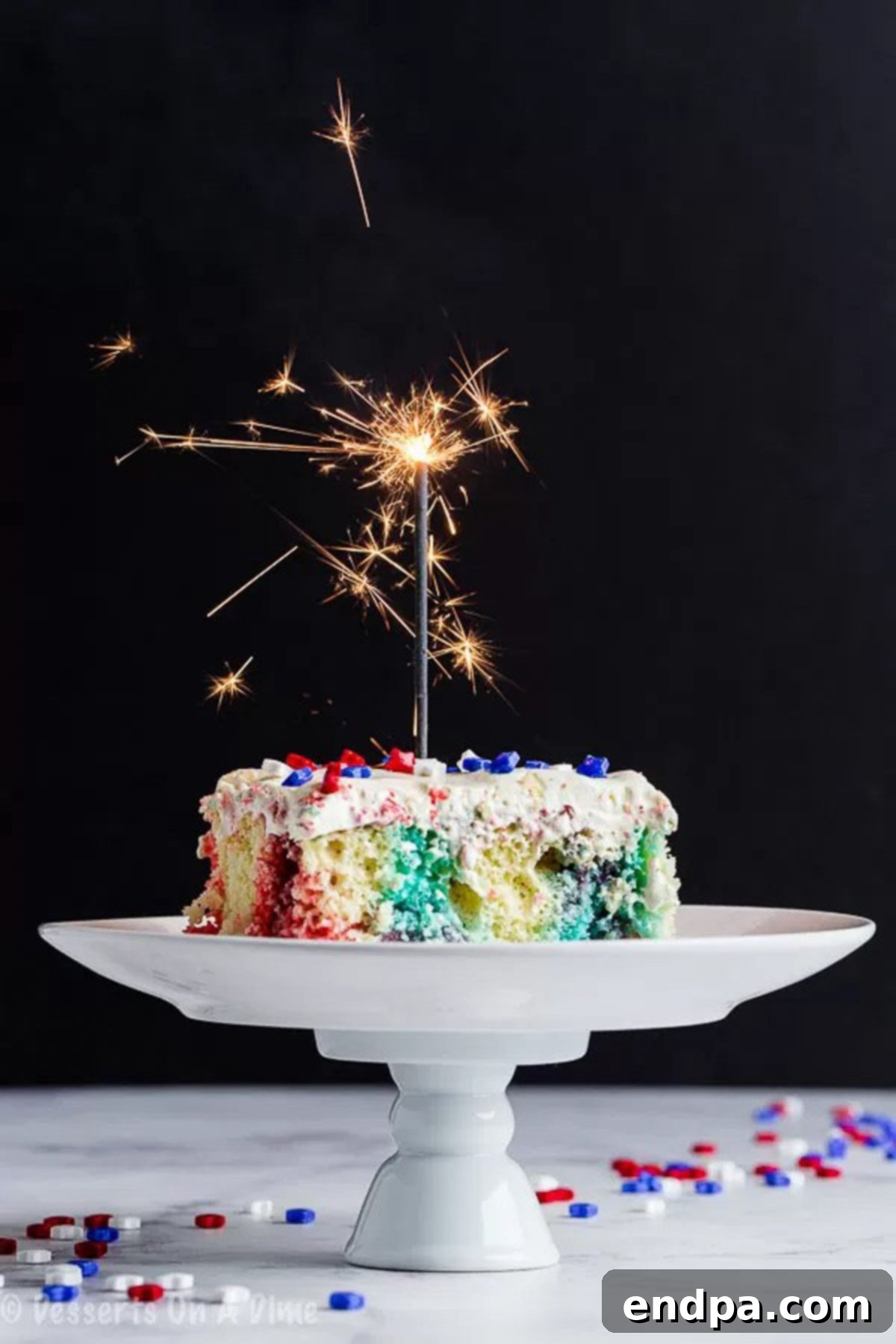 Close up image of a slice of patriotic poke cake on a white cake stand, with a small decorative firework in the background.