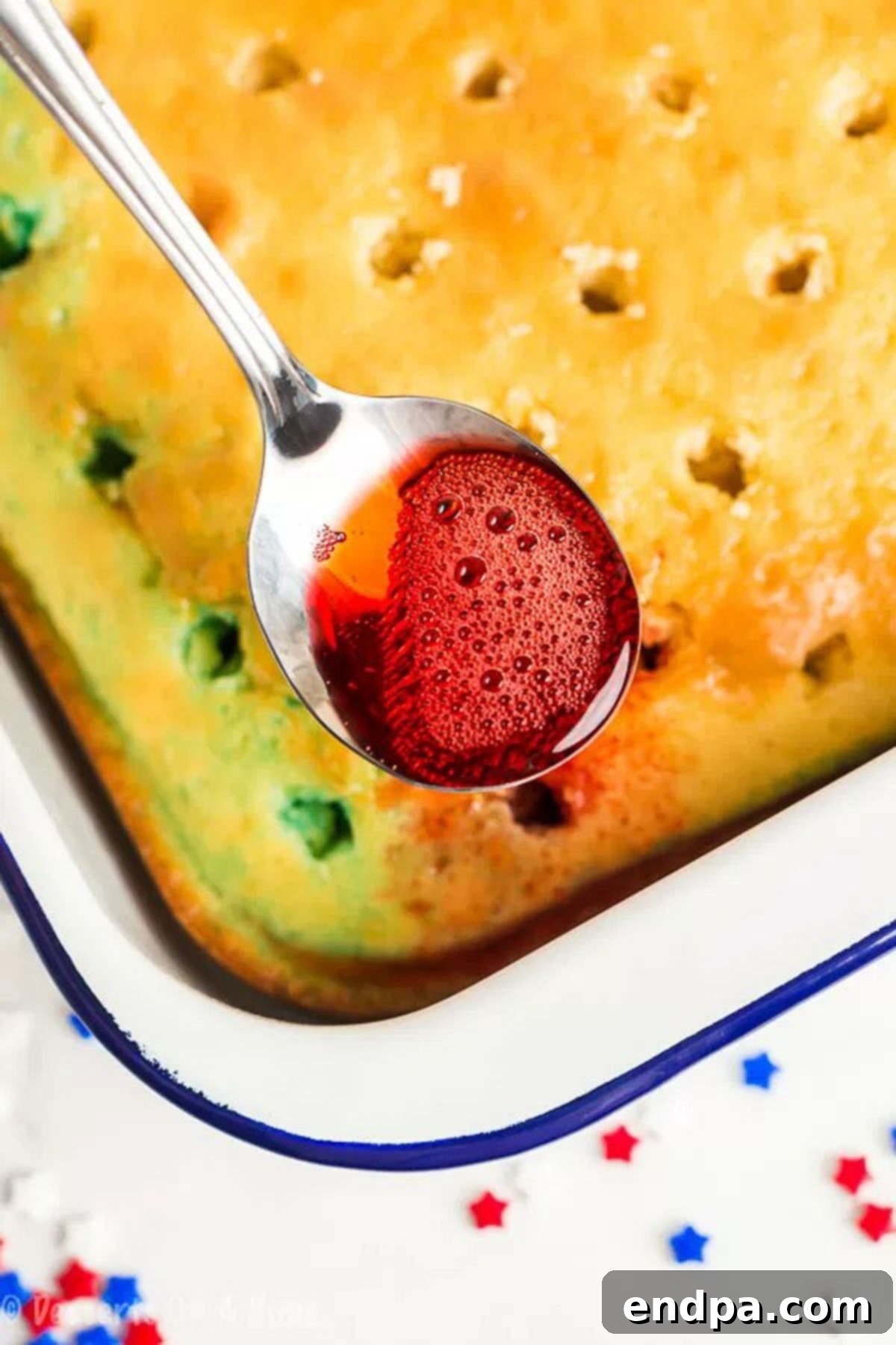 Close up image of a spoon of red Jell-O being poured over the poked cake.
