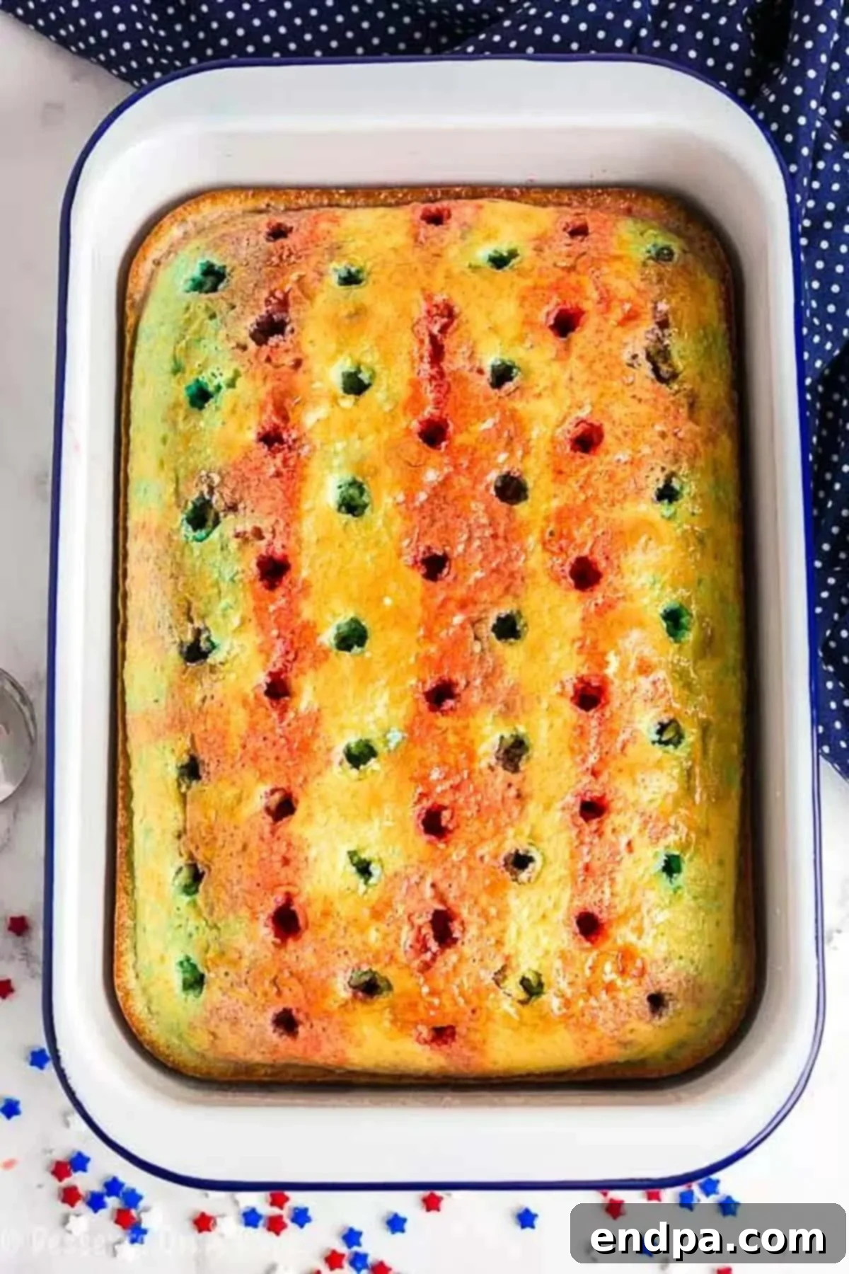 A close up image of a cake that has holes poked in it with red and blue Jell-O poured over it, chilling in the refrigerator.
