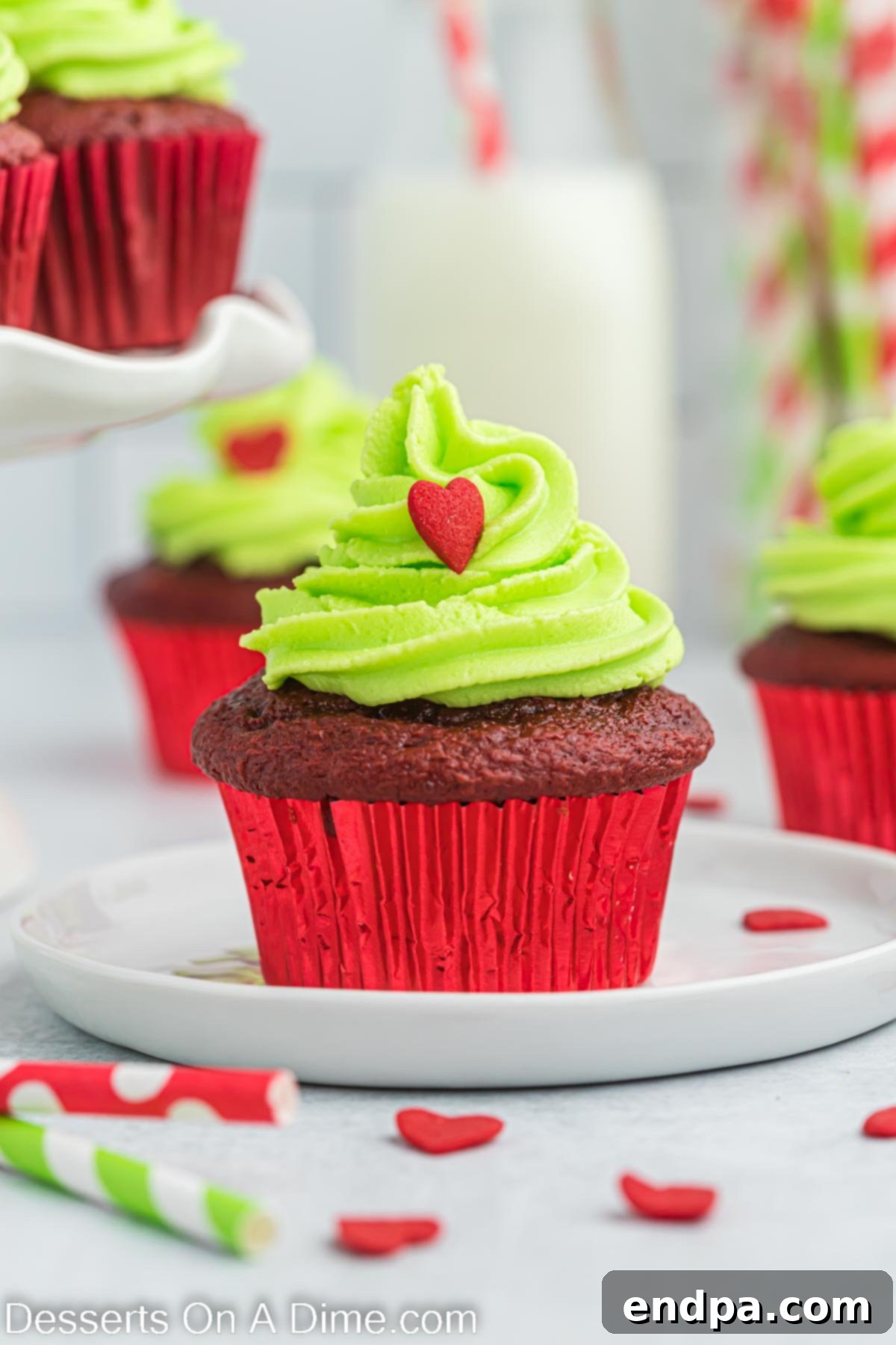 Close-up image of beautifully decorated Grinch cupcakes on a white plate, showcasing vibrant green frosting and tiny red heart sprinkles, ready for a holiday celebration.