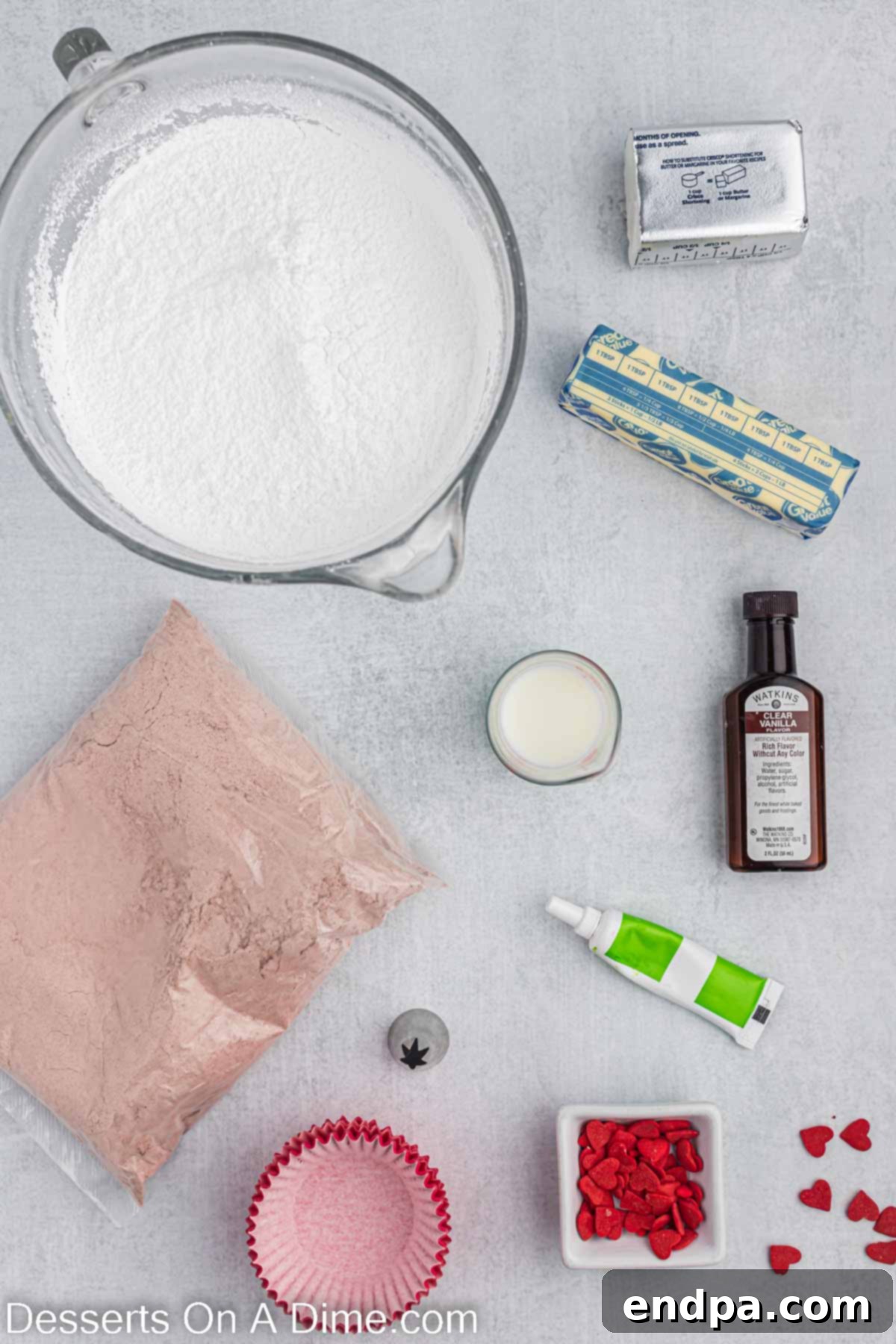 A flat lay photograph showcasing all the ingredients required for Grinch cupcakes: a red velvet cake mix box, bright red cupcake liners, softened butter, solid vegetable shortening, clear vanilla extract, a bag of powdered sugar, fresh milk, concentrated green gel food coloring, and charming red heart sprinkles.