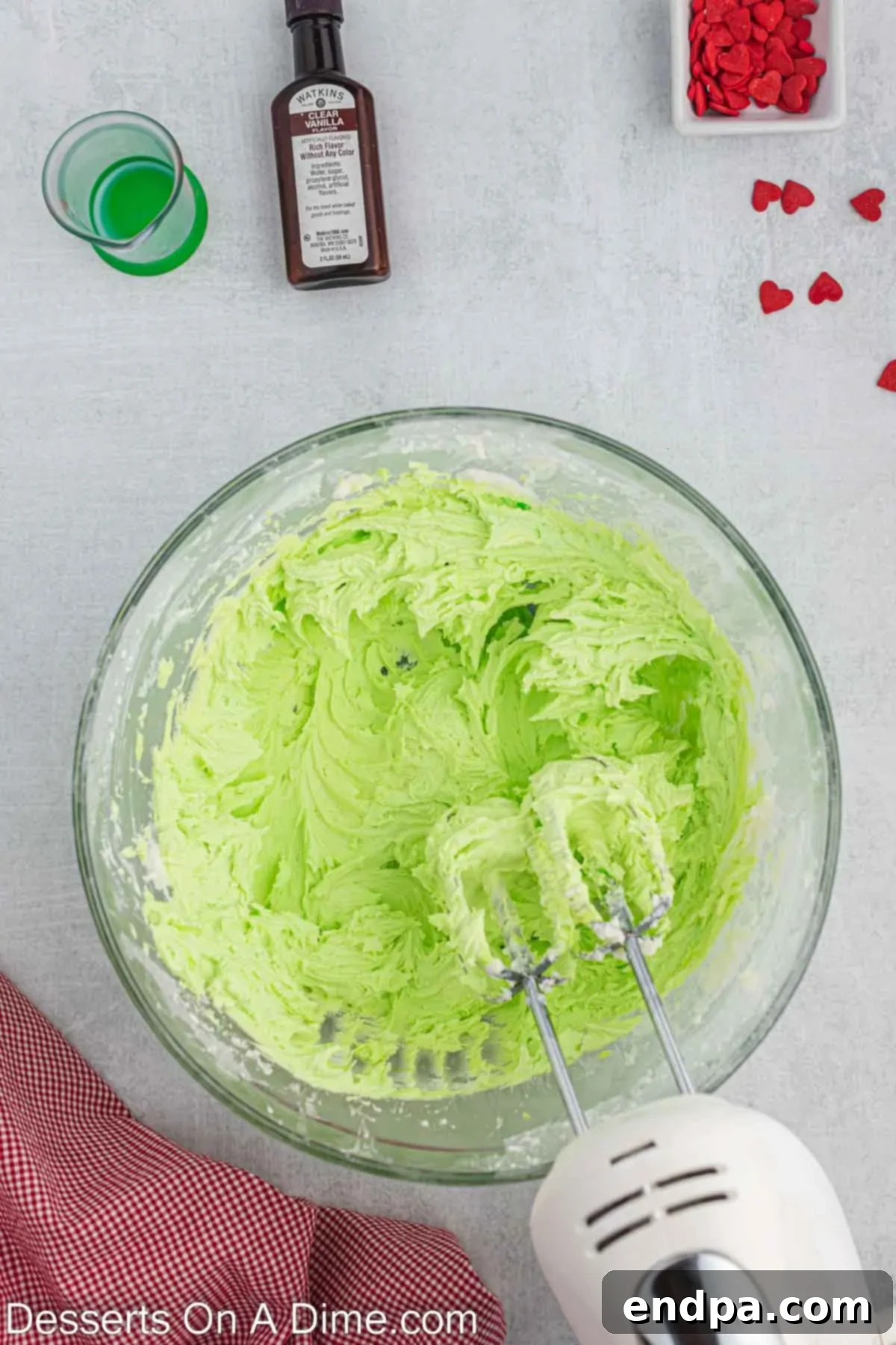 A close-up shot of vibrant green gel food coloring being carefully added drop by drop to the white buttercream frosting in a mixing bowl, to achieve the desired Grinch green hue.