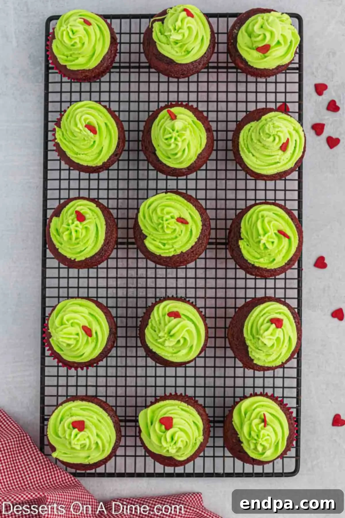 Grinch cupcakes on a wire rack, freshly frosted with a beautiful swirl of green icing and meticulously topped with a single red heart sprinkle, ready for the holidays.