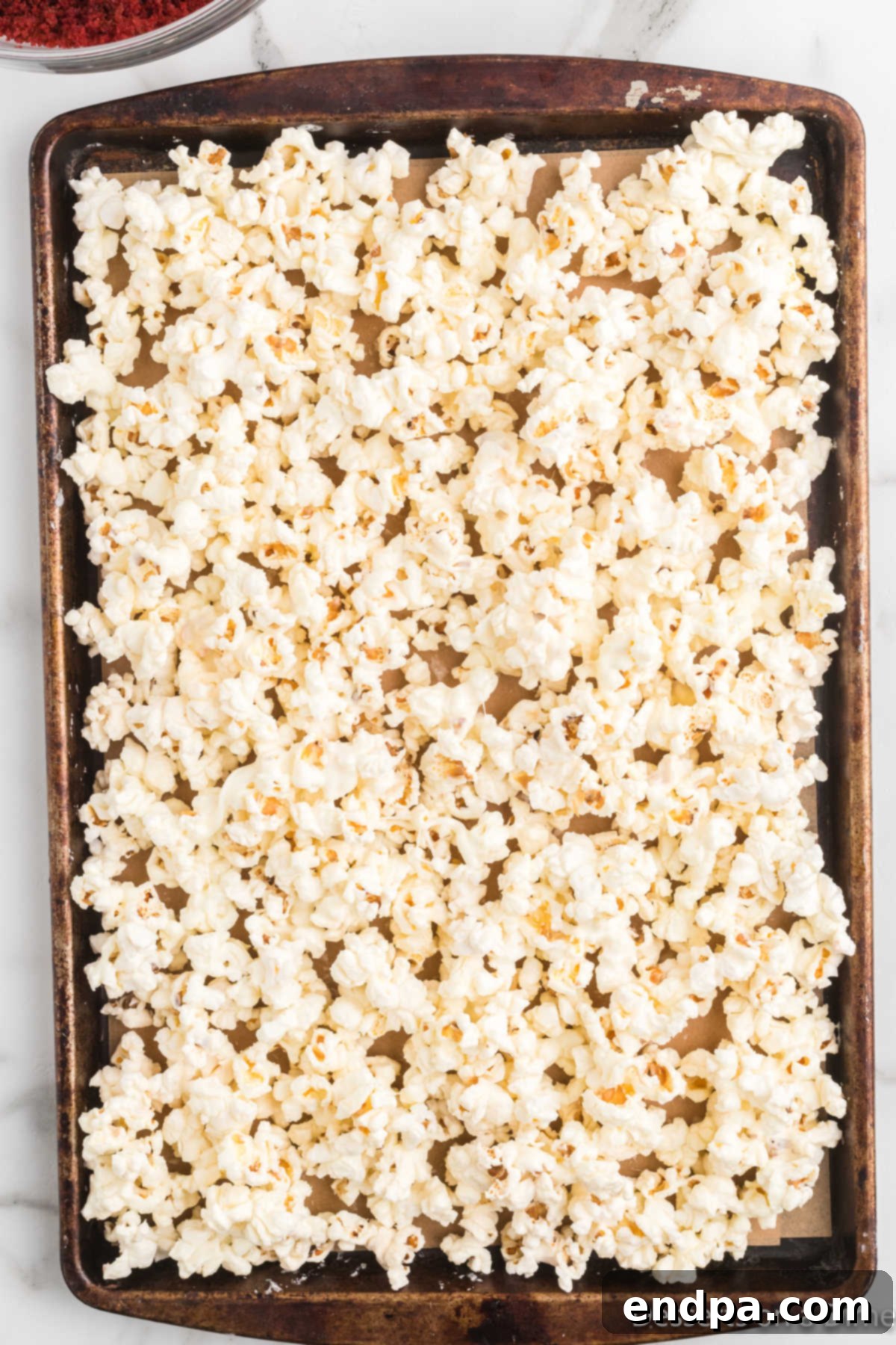 Popped popcorn spread evenly across a baking sheet lined with parchment paper, awaiting chocolate drizzle.