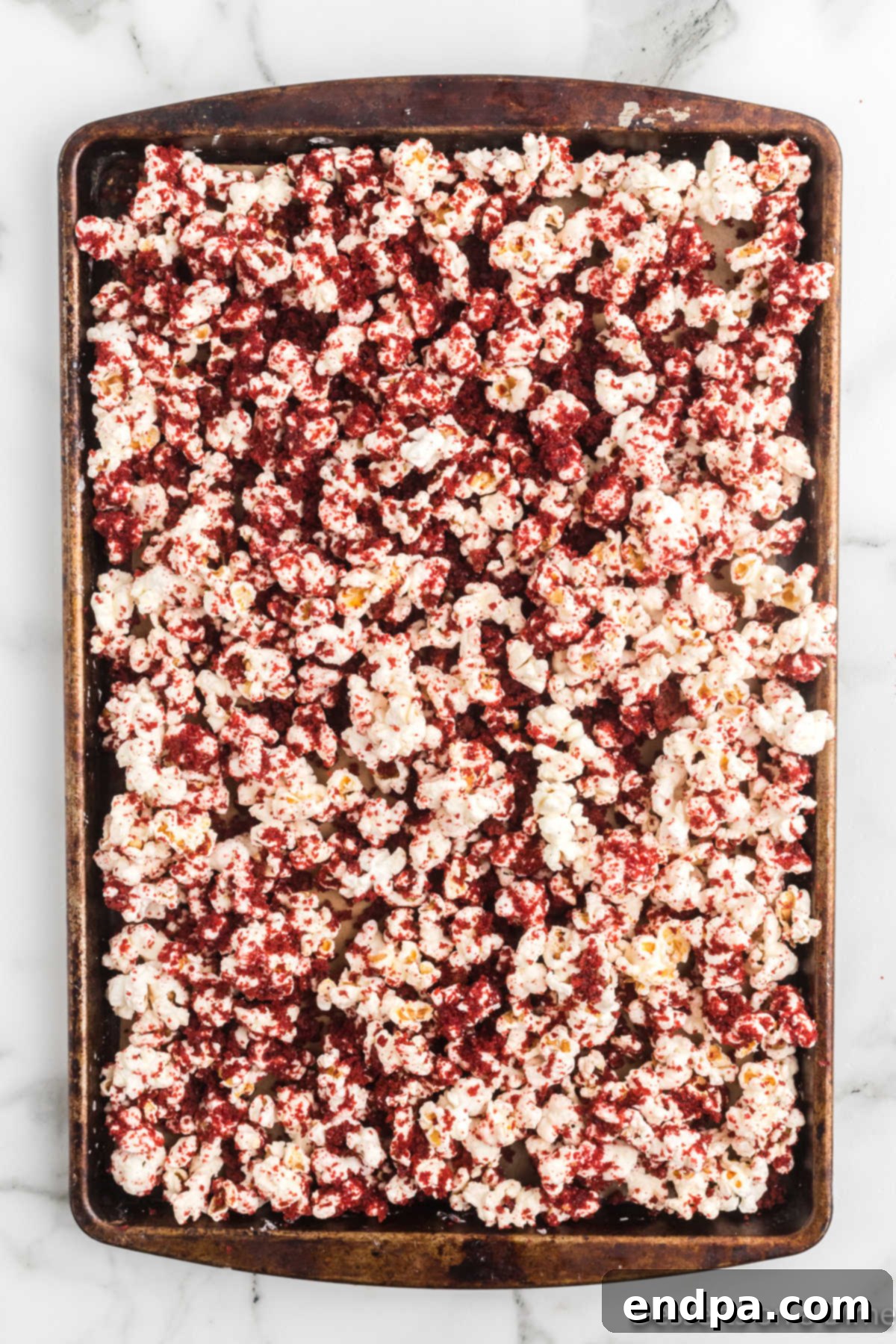 Red velvet cake crumbs being sprinkled generously over chocolate-coated popcorn on a baking sheet.
