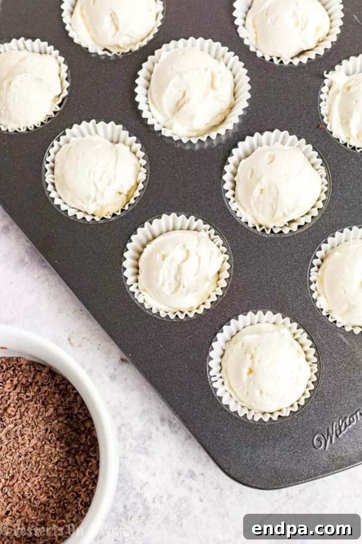 A mini muffin pan filled with Keto Cheesecake Fat Bombs in cupcake holders, positioned next to a small bowl of finely shaved sugar-free chocolate for topping, creating an enticing visual.