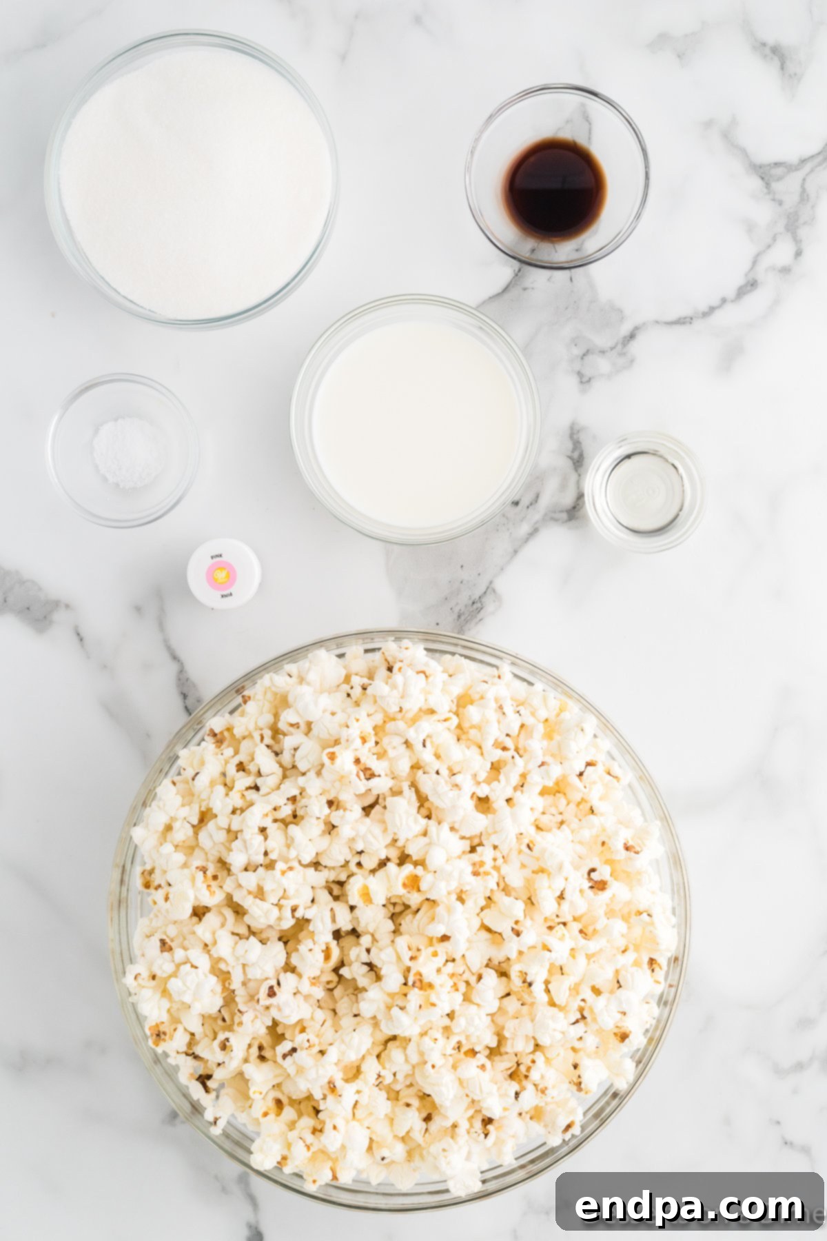 Ingredients for Pink Popcorn including popped popcorn, whole milk, vanilla extract, and pink food coloring, laid out on a kitchen surface.