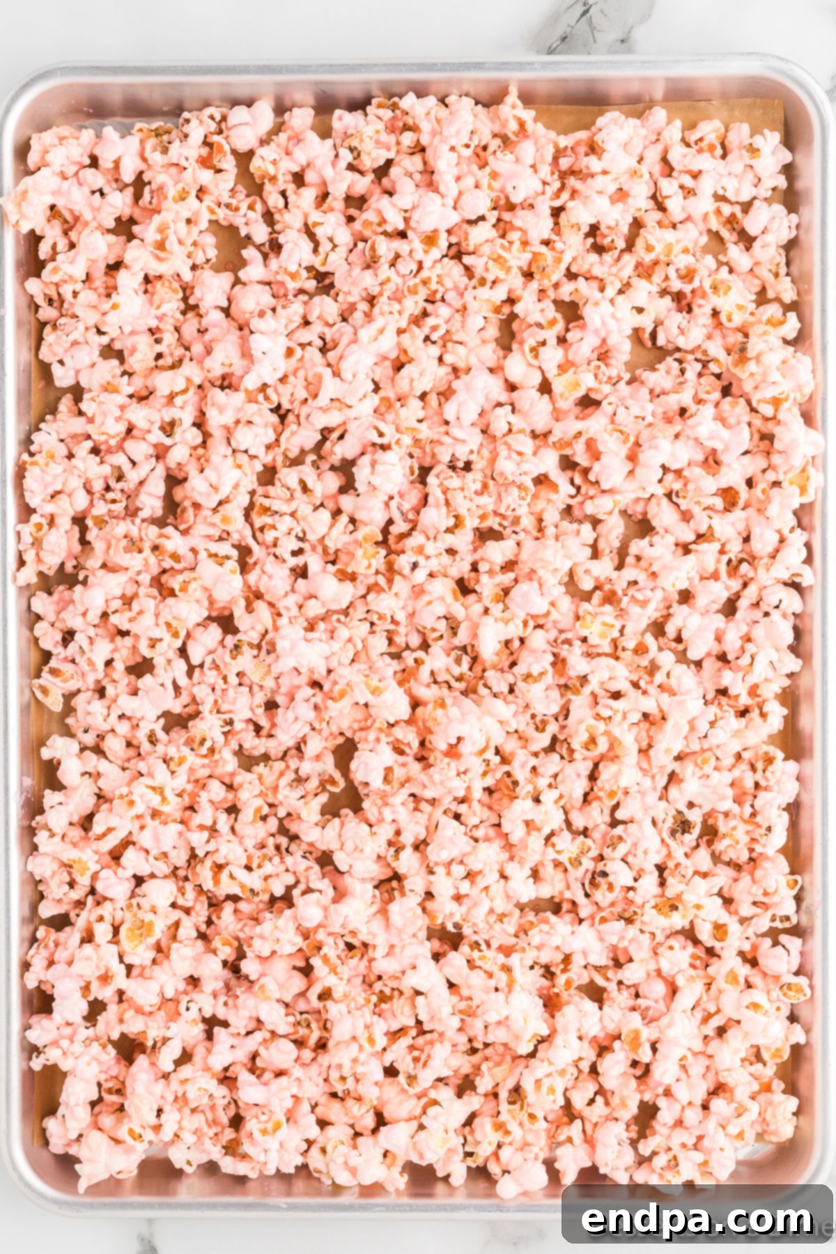 The pink sugar mixture spread over popped popcorn in a large bowl, being stirred to ensure even coating.
