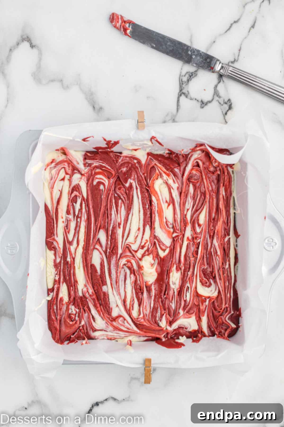 A knife gently swirling red and white fudge in a baking dish, creating beautiful marble patterns.