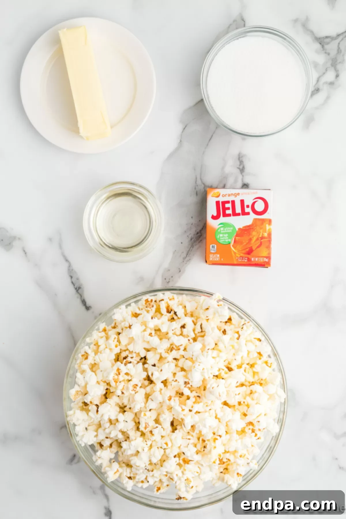 Rainbow Jello Popcorn Delight 3 Ingredients for Jello Popcorn - popcorn, butter, sugar, corn syrup, jello. A clear glass bowl holds freshly popped popcorn, while a stick of butter, a cup of granulated sugar, a bottle of light corn syrup, and a box of cherry Jello mix are neatly arranged around it. All items are clearly visible against a light background, emphasizing the simplicity of the recipe's components.