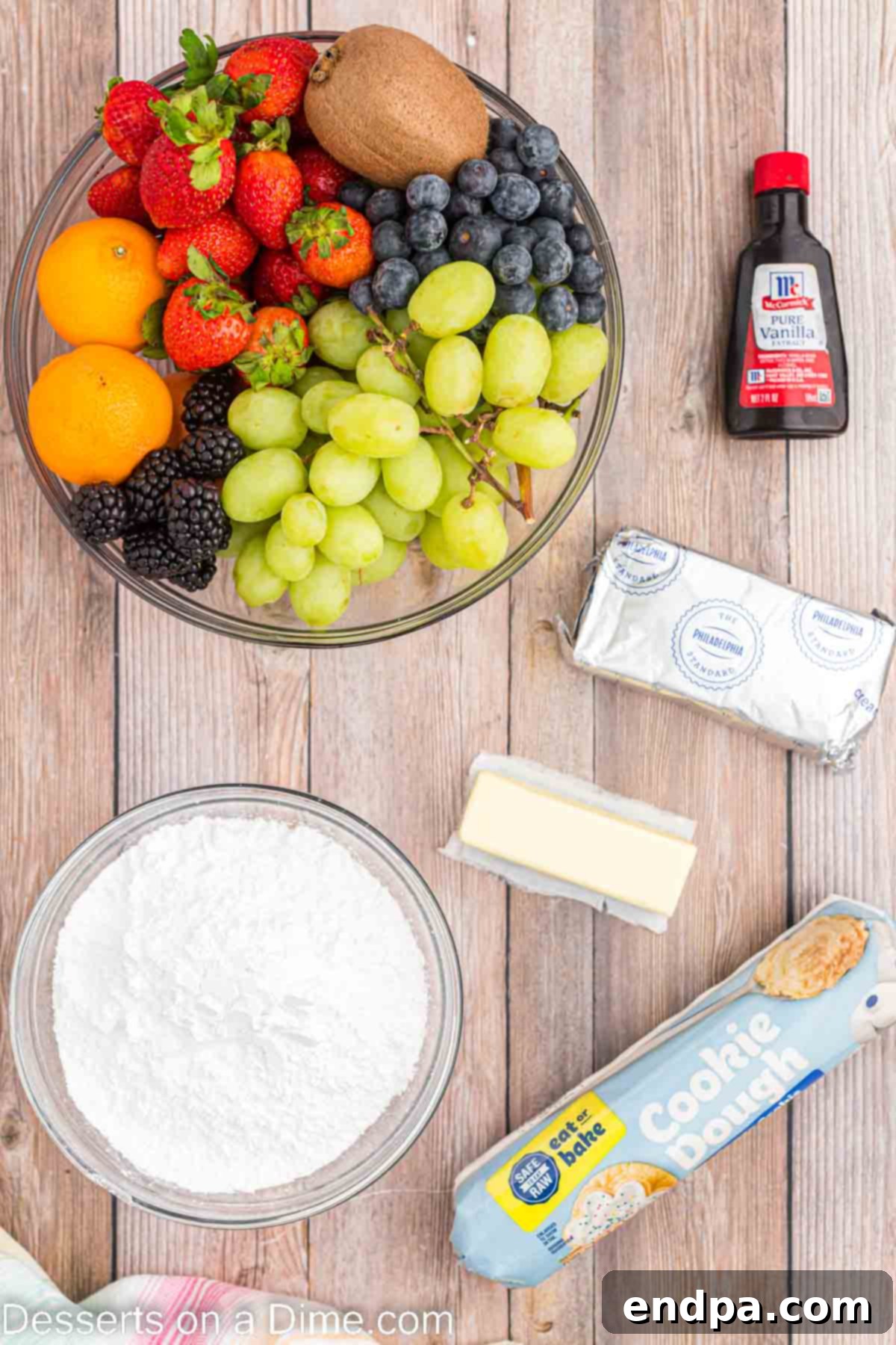 A collection of fresh ingredients laid out, including sugar cookie dough, cream cheese, butter, powdered sugar, vanilla extract, and assorted colorful fruits like strawberries, blueberries, and kiwis.