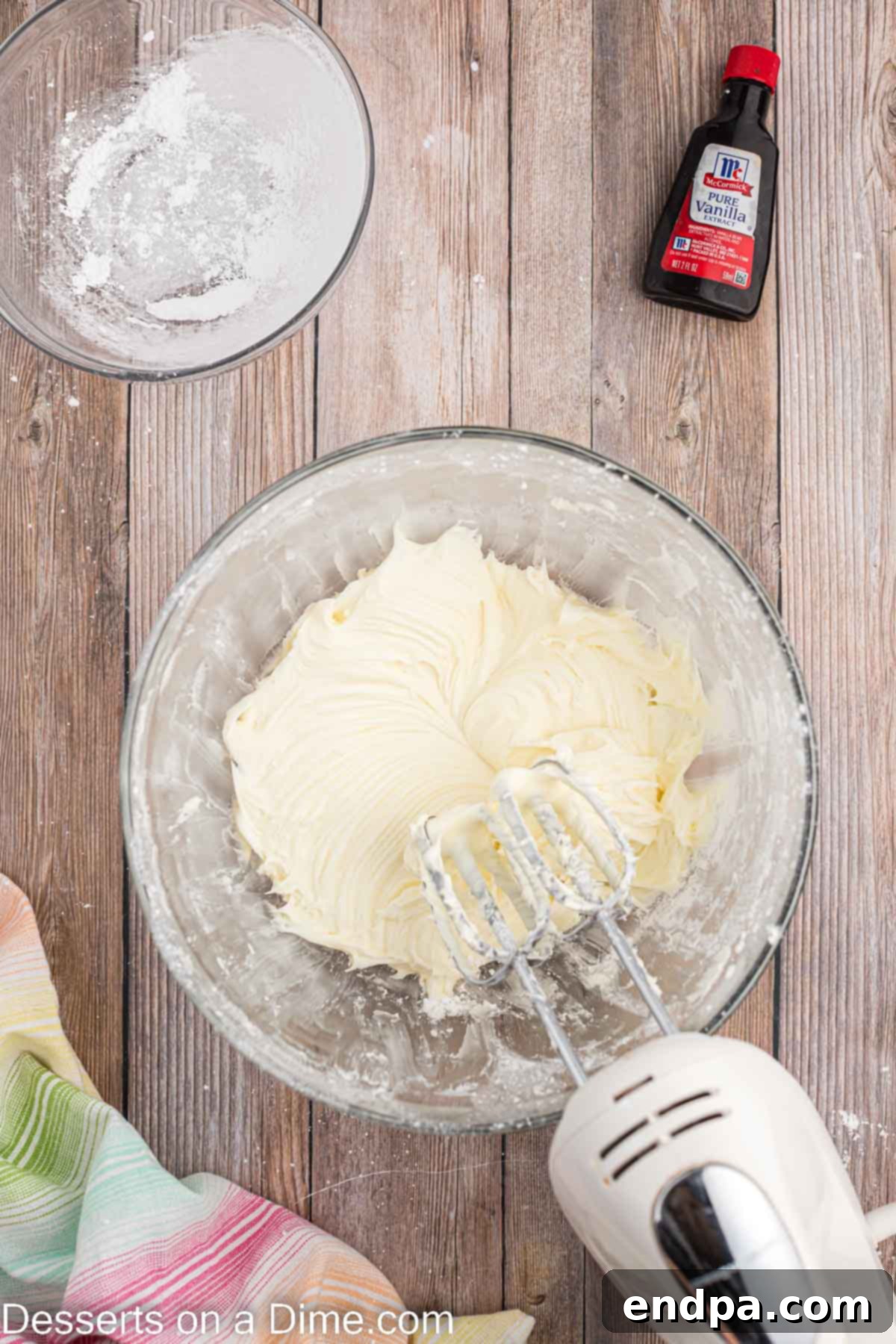 A mixing bowl filled with cream cheese, butter, powdered sugar, and vanilla extract, being blended with an electric hand mixer.