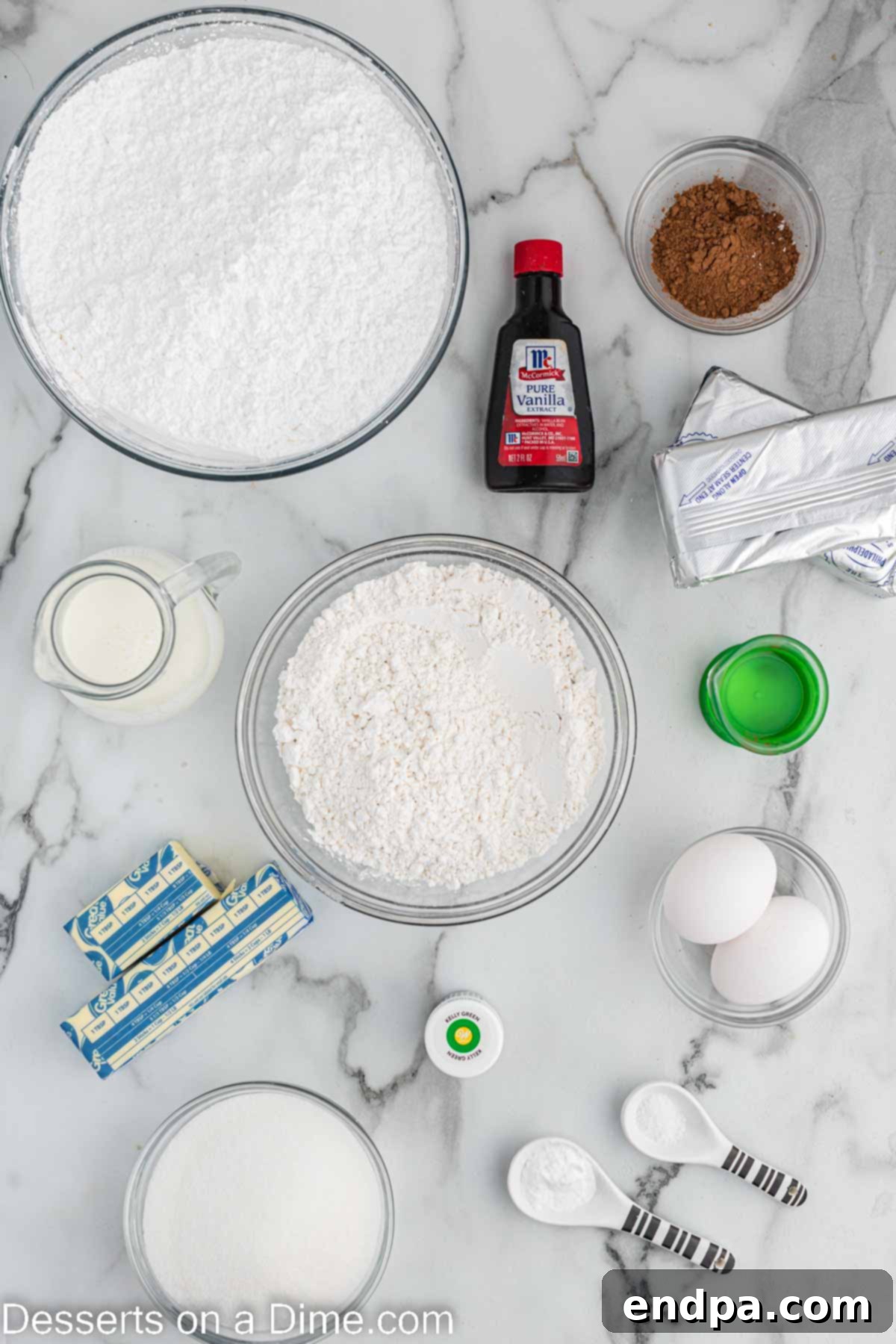 A collection of ingredients laid out on a table for making green velvet cupcakes, including cake flour, cocoa powder, sugar, oil, green gel food coloring, buttermilk, cream cheese, butter, and powdered sugar.