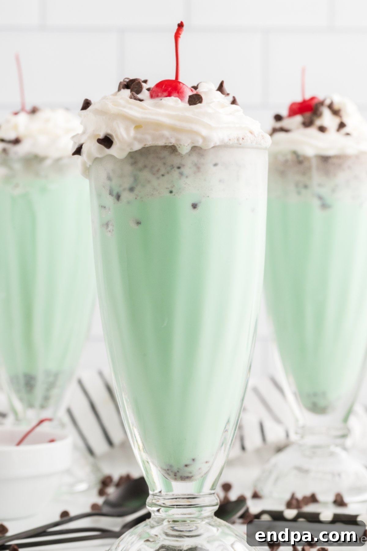 Two glasses of vibrant green mint chocolate chip milkshakes, topped with whipped cream, chocolate chips, and a cherry, against a blurred background.