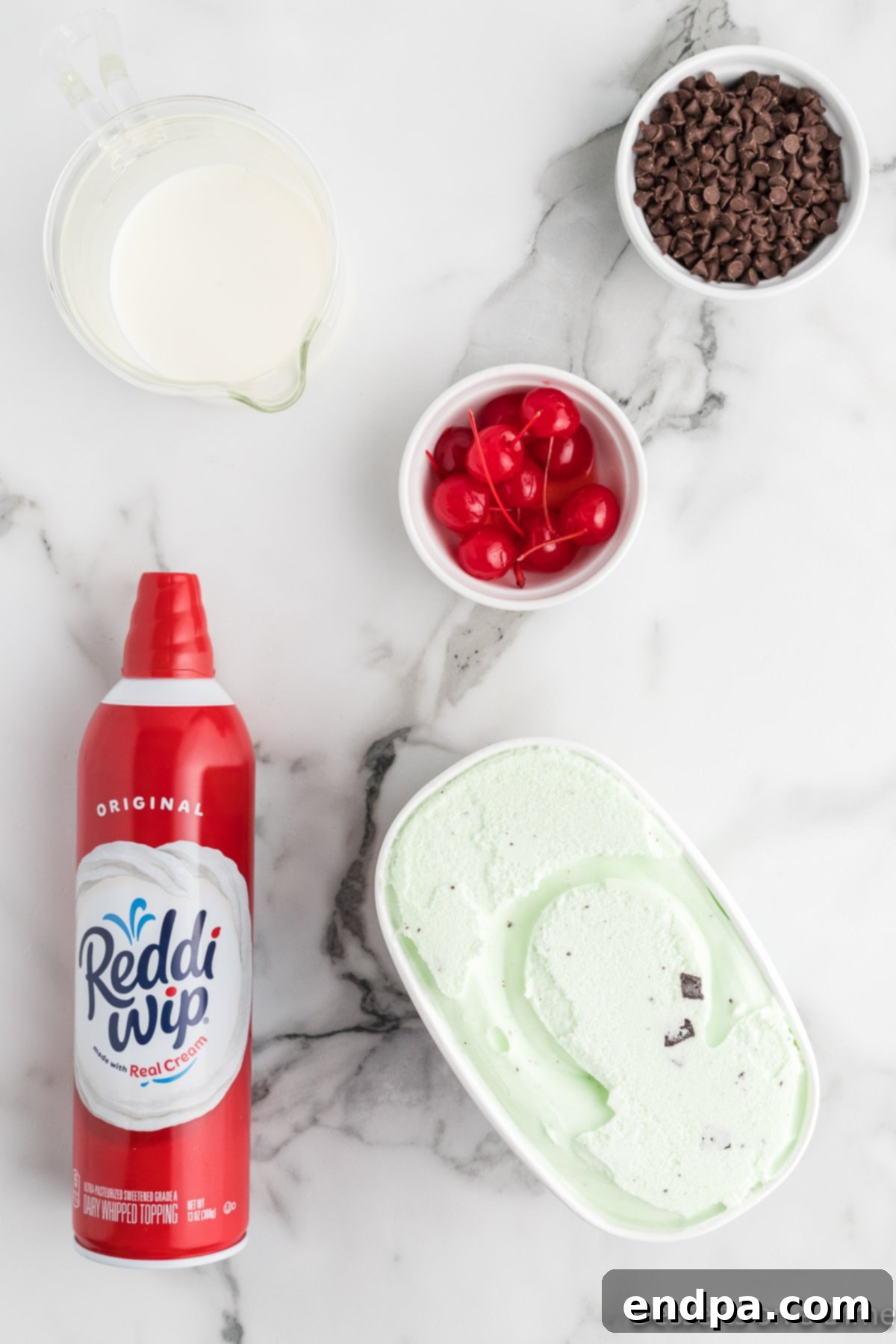A flat lay image showcasing the simple ingredients for mint chocolate chip milkshakes: a carton of mint chocolate chip ice cream, a bottle of whole milk, a can of whipped cream, a bowl of mini chocolate chips, and a jar of maraschino cherries.