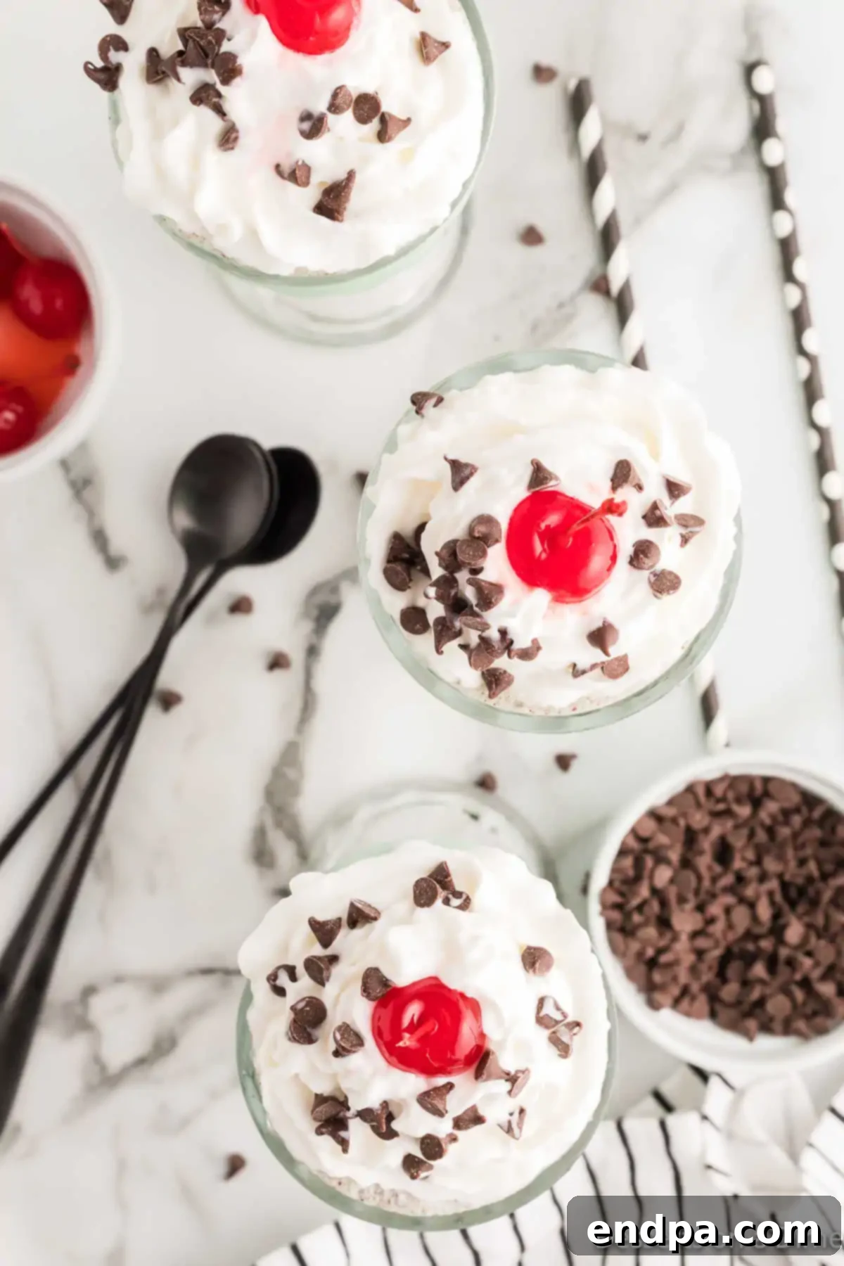 Two glasses of mint chocolate chip milkshakes, now topped generously with whipped cream, mini chocolate chips, and a single maraschino cherry on each.
