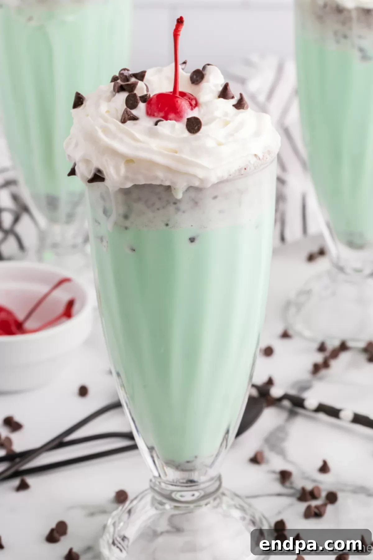 Close-up of two glasses of mint chocolate chip milkshakes, garnished with whipped cream, chocolate chips, and cherries, ready to be enjoyed.