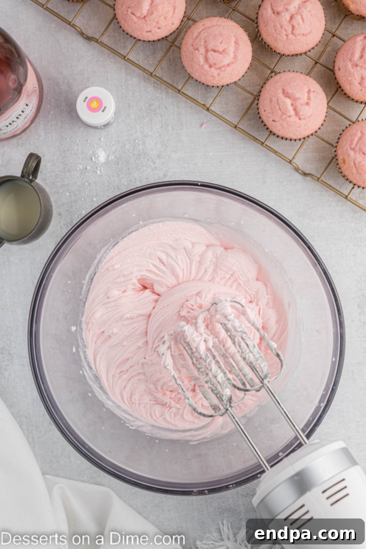 Pink food coloring being added to the white buttercream frosting in a bowl, with a splash of milk.