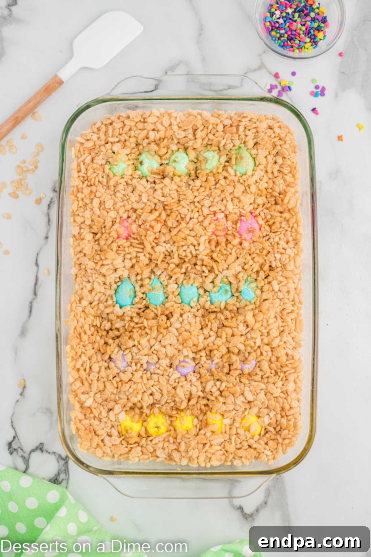 The melted Peeps, butter, and cereal mixture being stirred and smoothed out in the baking dish with a spatula.