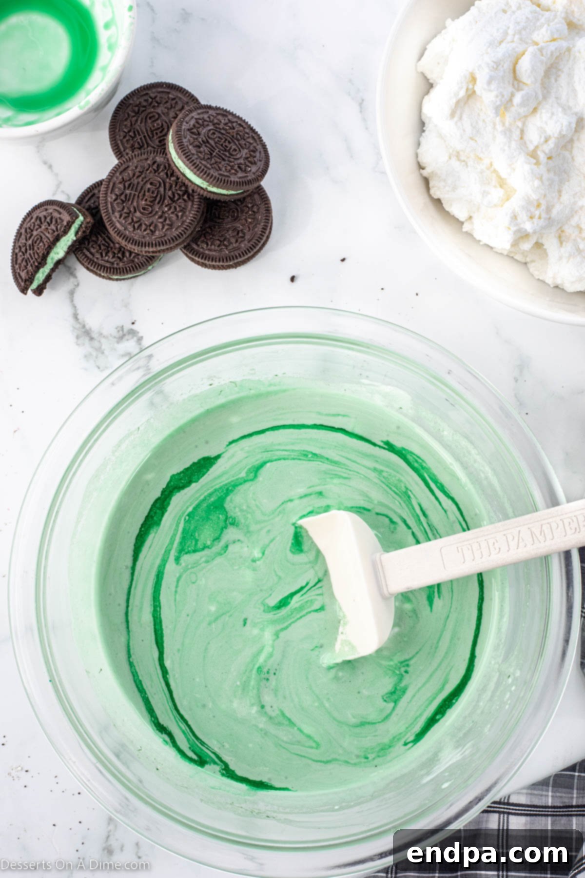 Emerald Mint Delight 7 A mixing bowl containing marshmallow creme, green food coloring, Crème de Menthe, and Crème de Cacao, being combined with a spatula.