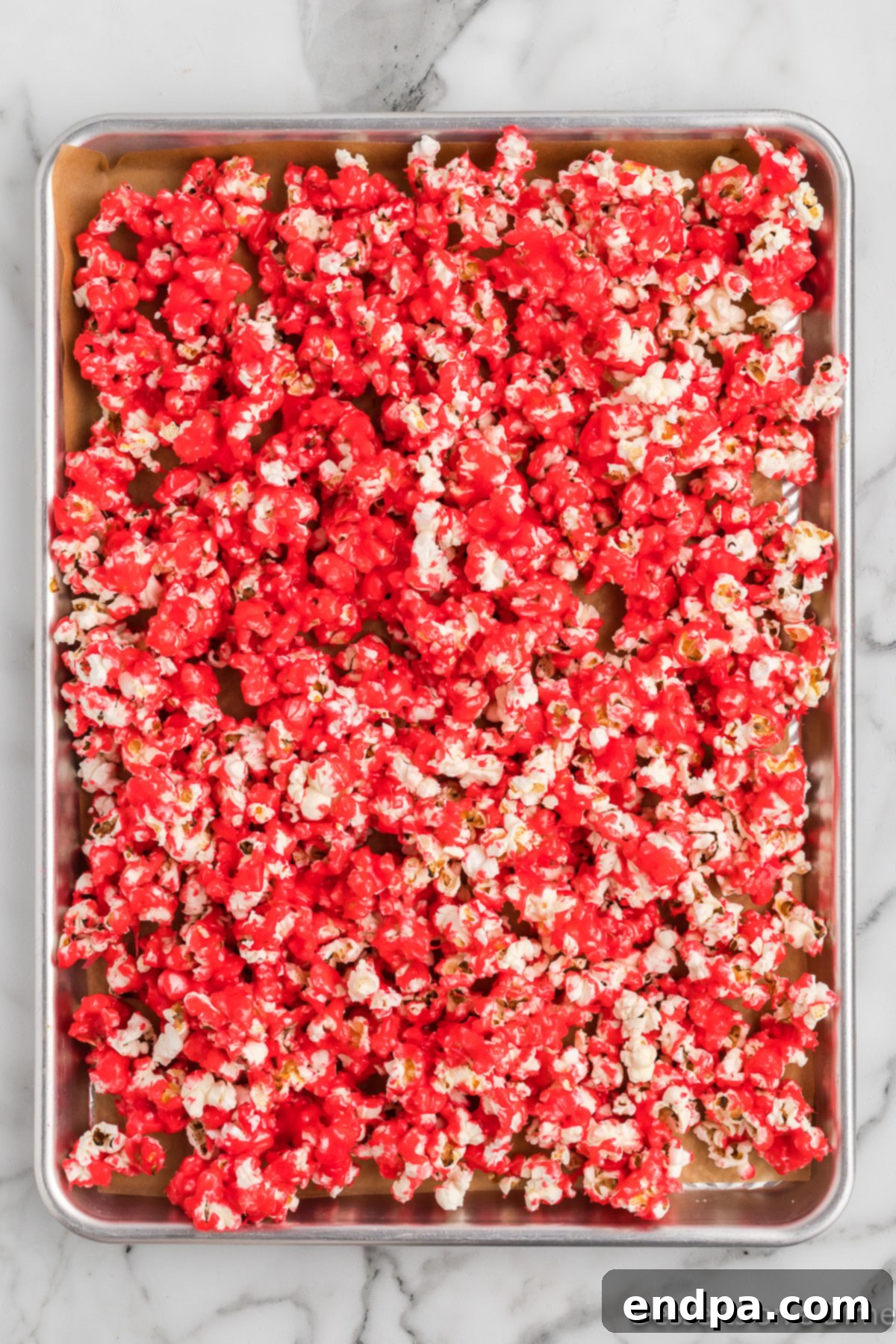 Red Hot Cinnamon Popcorn spread out on a sheet pan, glistening with its candy coating, ready for baking to achieve maximum crispness.
