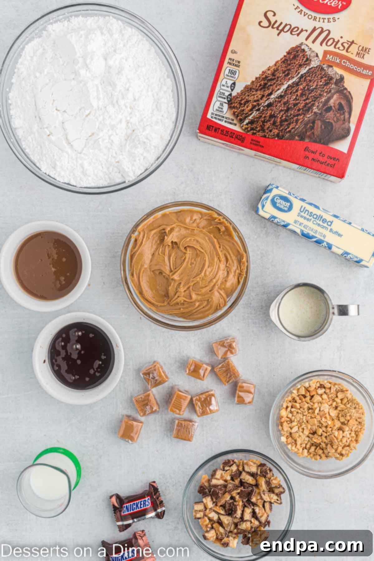 Ingredients for cupcakes - cake mix, caramel candy, heavy cream, salted peanuts, butter, peanut butter, powdered sugar, caramel sauce, chocoalte sauce, snicker bars, laid out on a white surface.