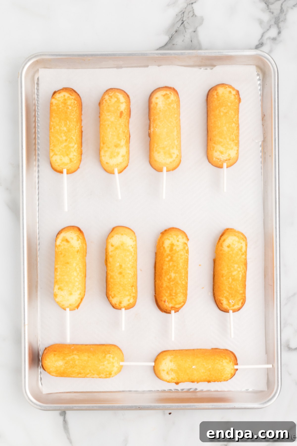 Decadent Chocolate Dipped Twinkies 4 Unwrapped Twinkies with lollipop sticks inserted, placed neatly on a baking sheet lined with parchment paper.