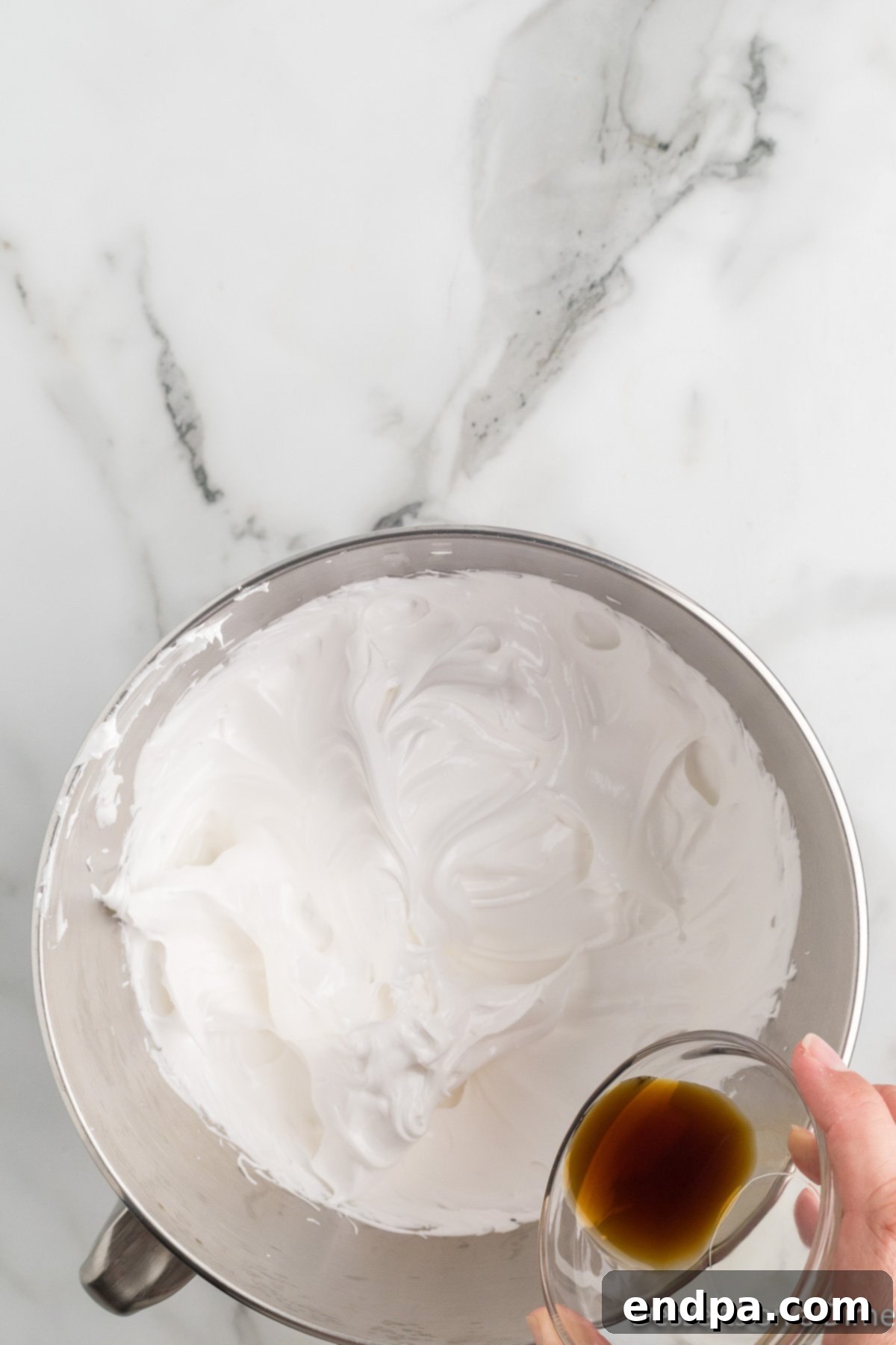 Vanilla extract being poured into the thick, glossy marshmallow mixture during the final stages of whipping.