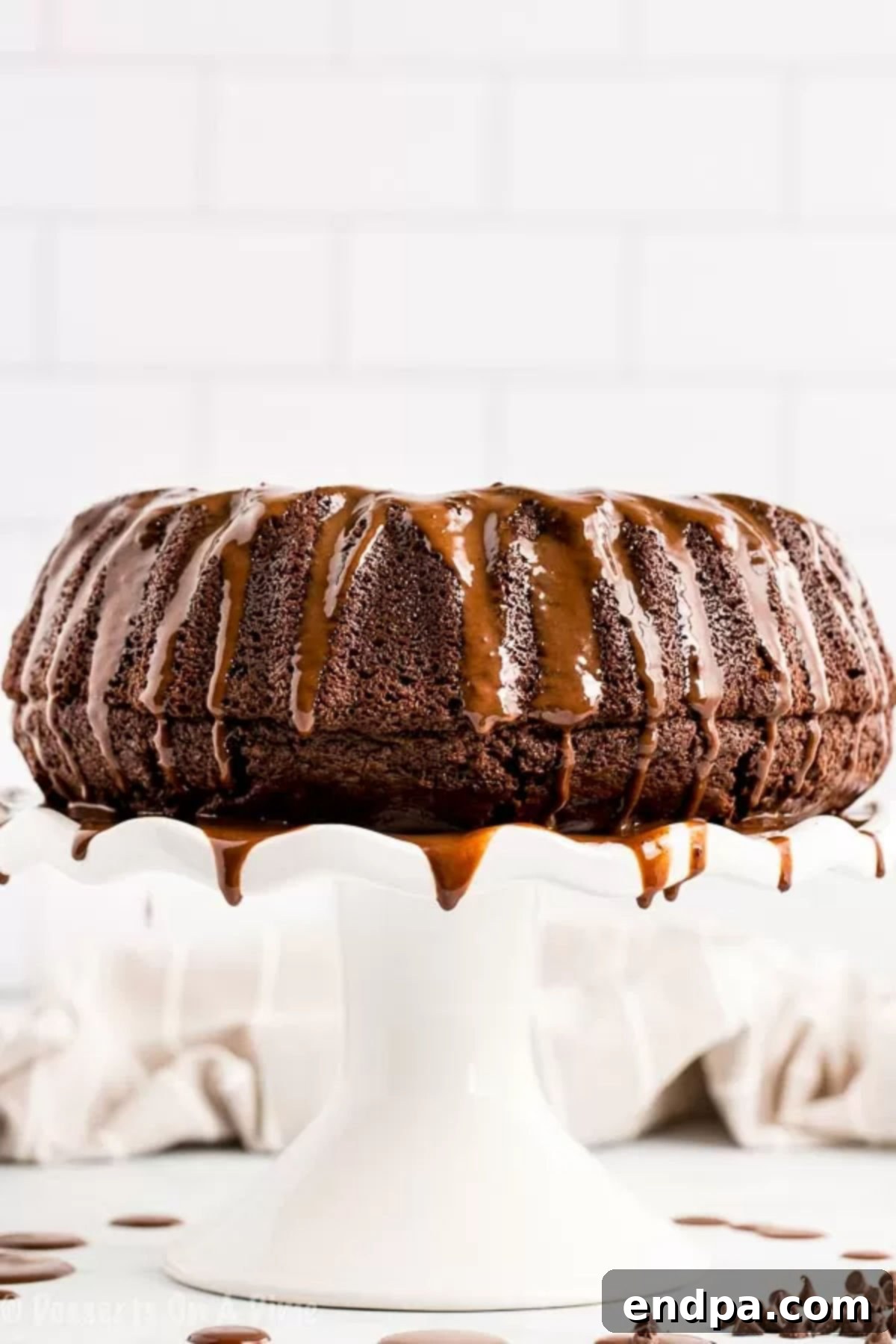 A stunning Chocolate Bundt Cake on a white cake stand, drizzled with rich chocolate glaze.