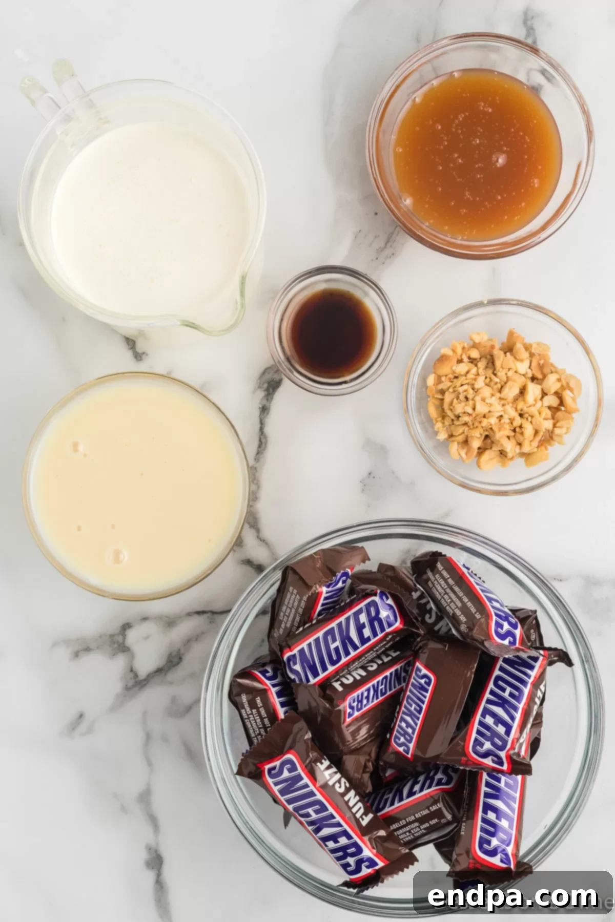 Decadent Homemade Snickers Ice Cream 3 A collection of ingredients laid out for Snickers ice cream, including a can of sweetened condensed milk, a bottle of vanilla extract, a carton of heavy whipping cream, a jar of caramel sauce, a bag of fun-size Snickers candy bars, and a small bowl of chopped peanuts.