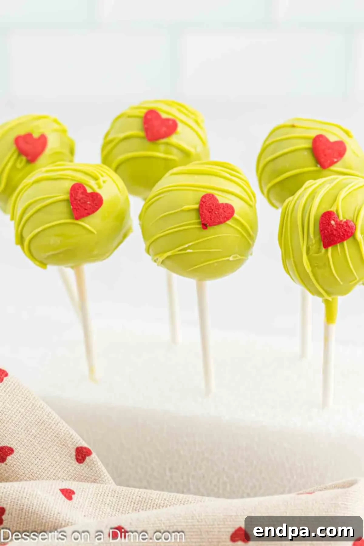 Grinchmas Cake Pops 2 Grinch Cake Pops on a festive stand, showcasing their vibrant green coating and red candy hearts.