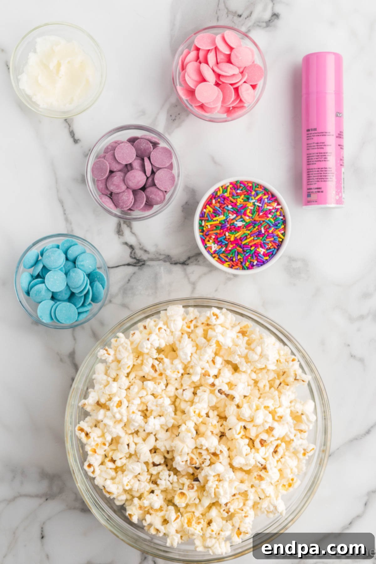 A flat lay image displaying the key ingredients for Unicorn Popcorn: a bowl of freshly popped popcorn, a can of pink candy spray, three separate bowls of pink, blue, and purple candy melts, and a jar of rainbow sprinkles.