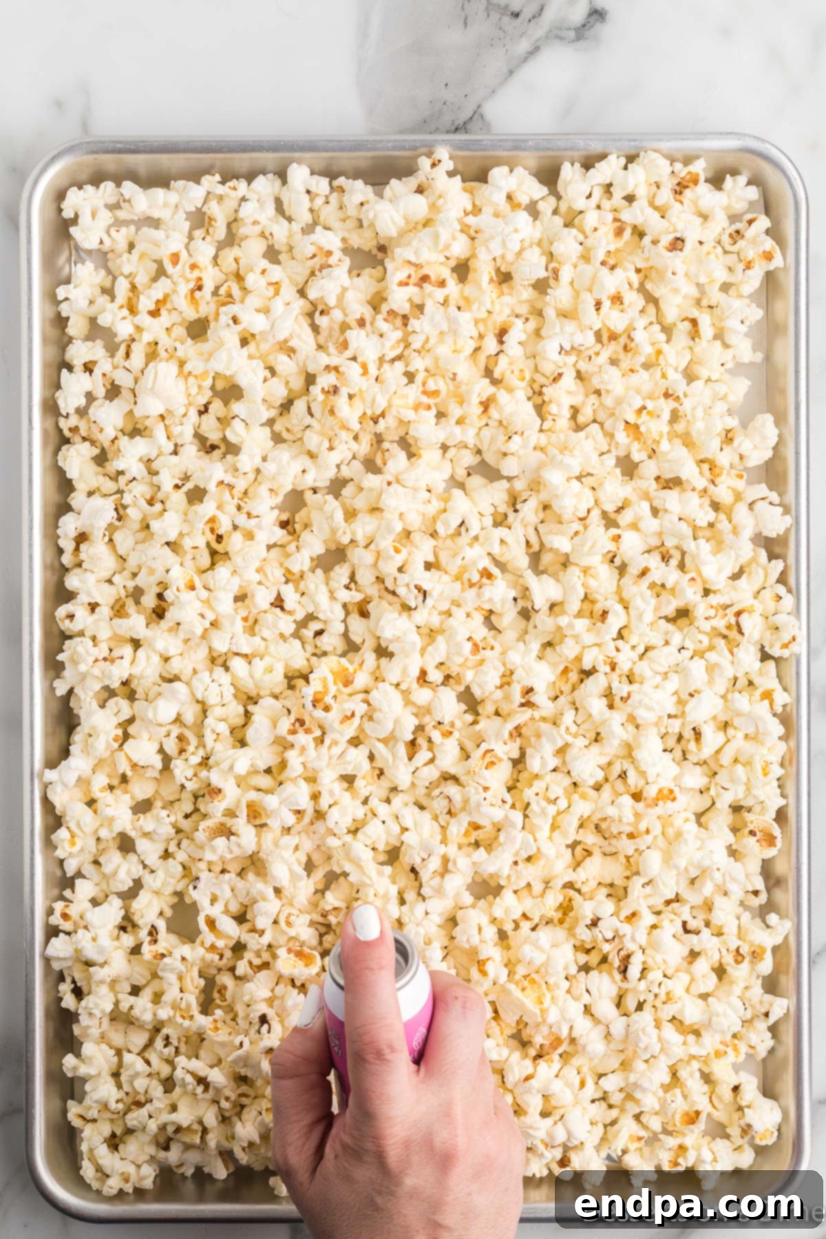 Freshly popped popcorn spread evenly across a baking sheet lined with parchment paper, preparing for the candy coating.