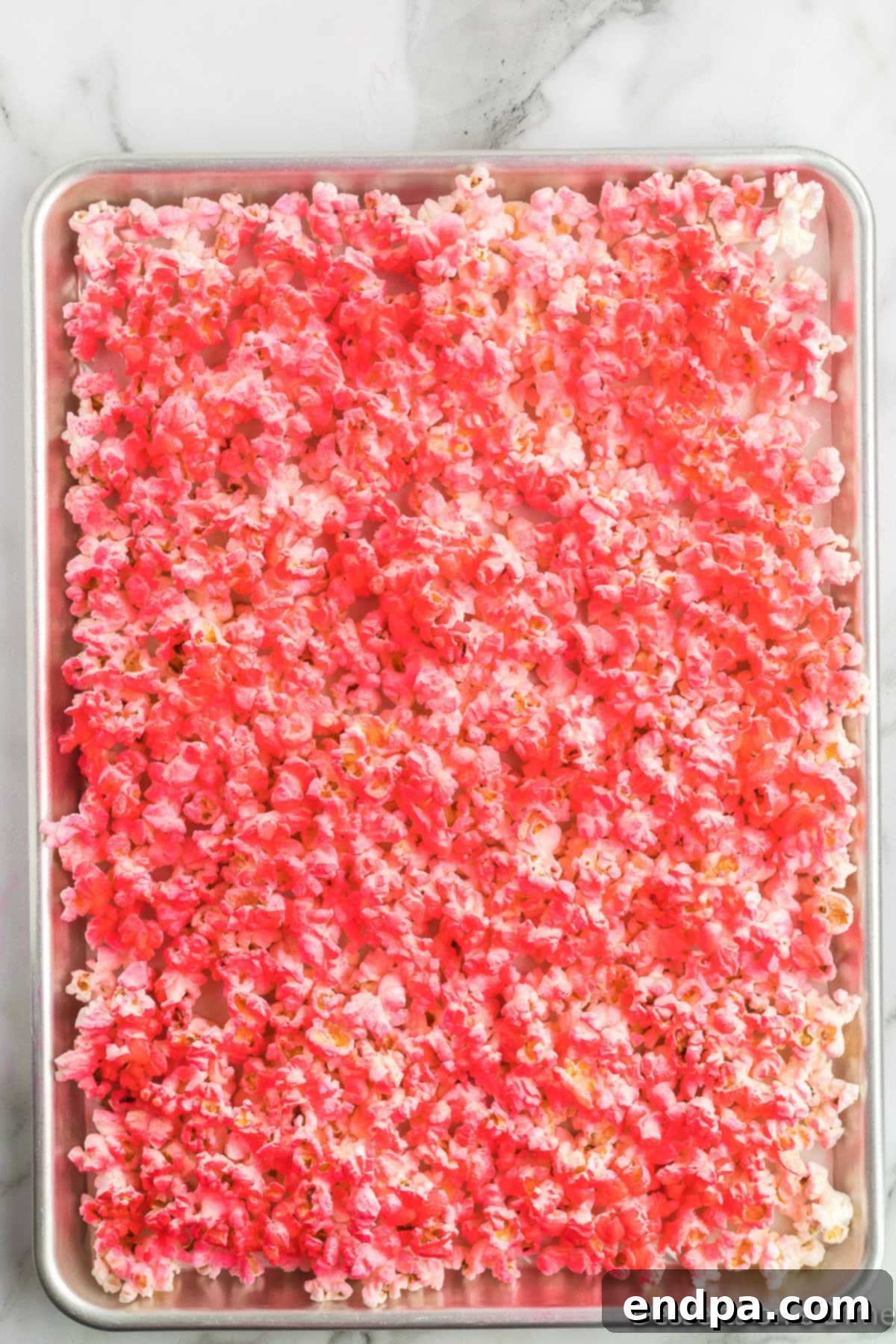 Popcorn on a baking sheet being sprayed with pink candy food spray, creating a vibrant base color before the candy melts are added.