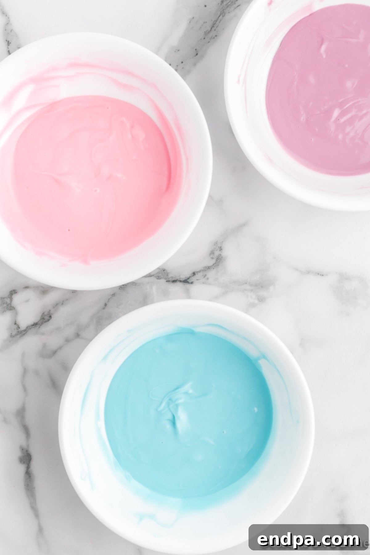 Three separate microwave-safe bowls containing melted pink, blue, and purple candy melts, each with a smooth, pourable consistency.