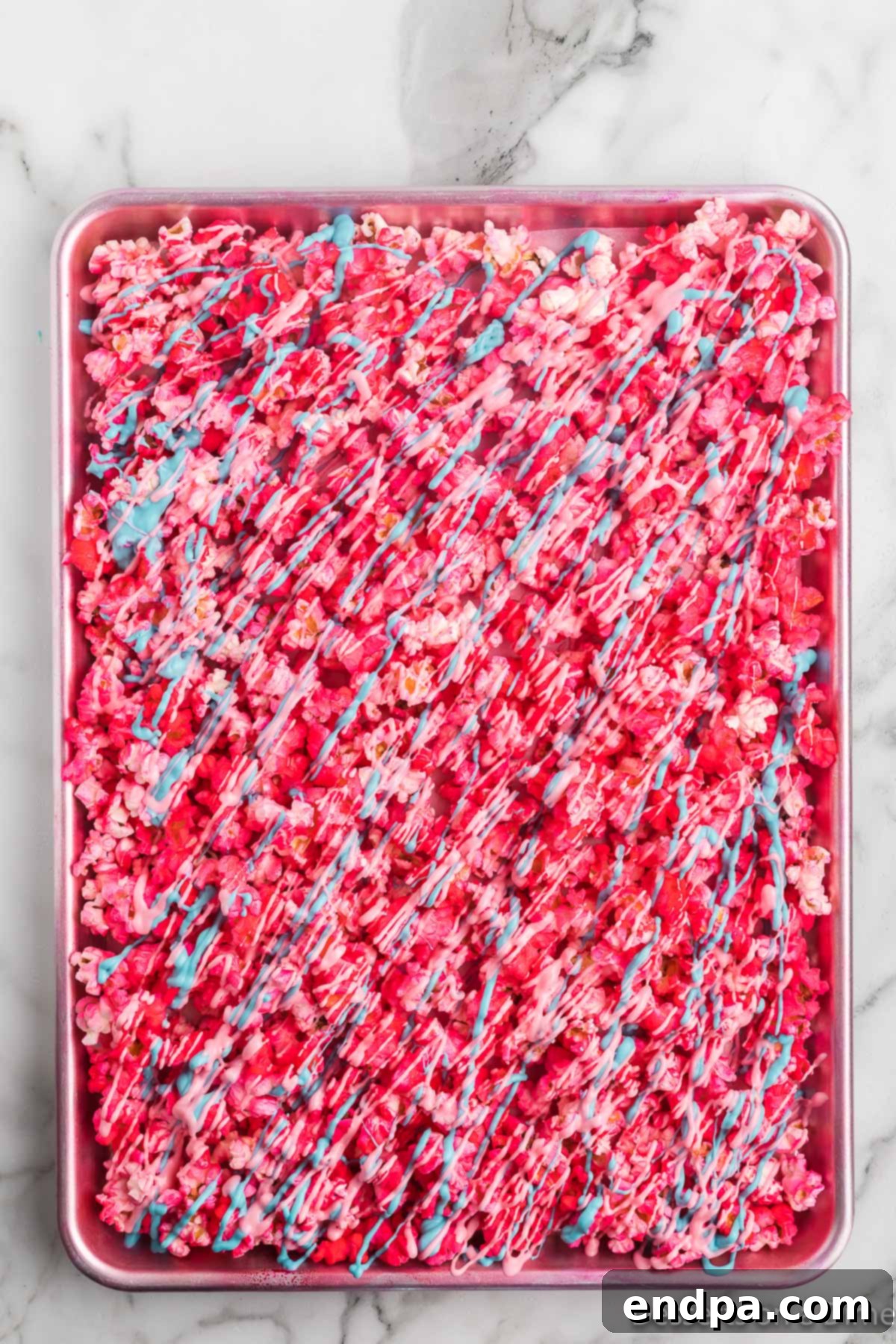 Melted candy melts in pink, blue, and purple being drizzled artistically over the prepared popcorn on a baking sheet, with rainbow sprinkles waiting to be added.