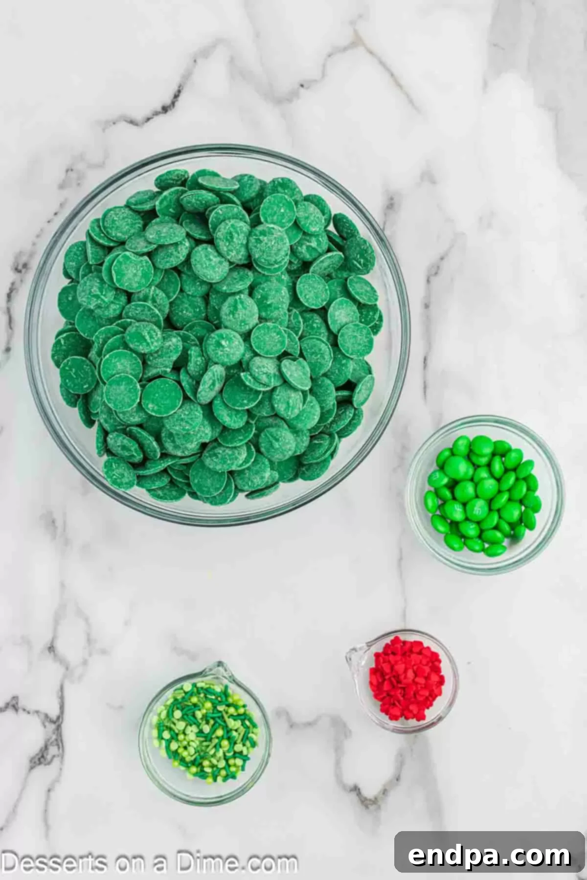 Ingredients for Grinch Bark: green candy melts, green M&Ms, green sprinkles, and red candy hearts displayed on a wooden surface.