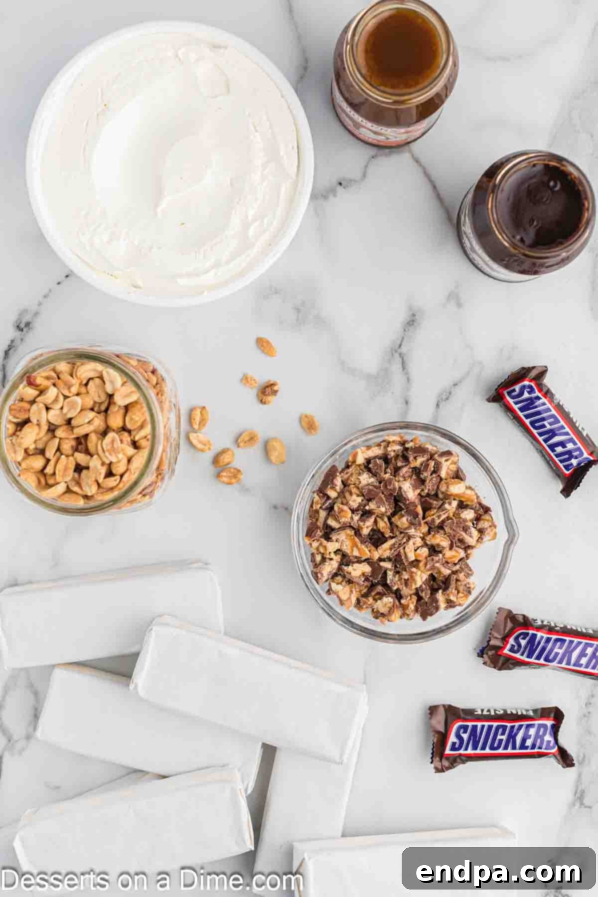 Ultimate Snickers Ice Cream Cake 3 A flat lay image showing all the ingredients required for Snickers ice cream cake: ice cream sandwiches, jars of hot fudge and caramel, a tub of whipped topping, a bowl of chopped Snickers bars, and a bowl of roasted salted peanuts.