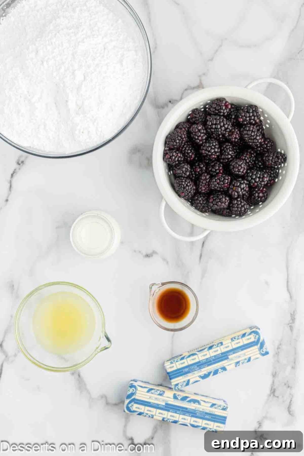 A flat lay showing fresh blackberries, a lemon, a stick of unsalted butter, a bowl of powdered sugar, a bottle of vanilla extract, and a small jug of milk – all essential ingredients for Blackberry Buttercream Frosting.