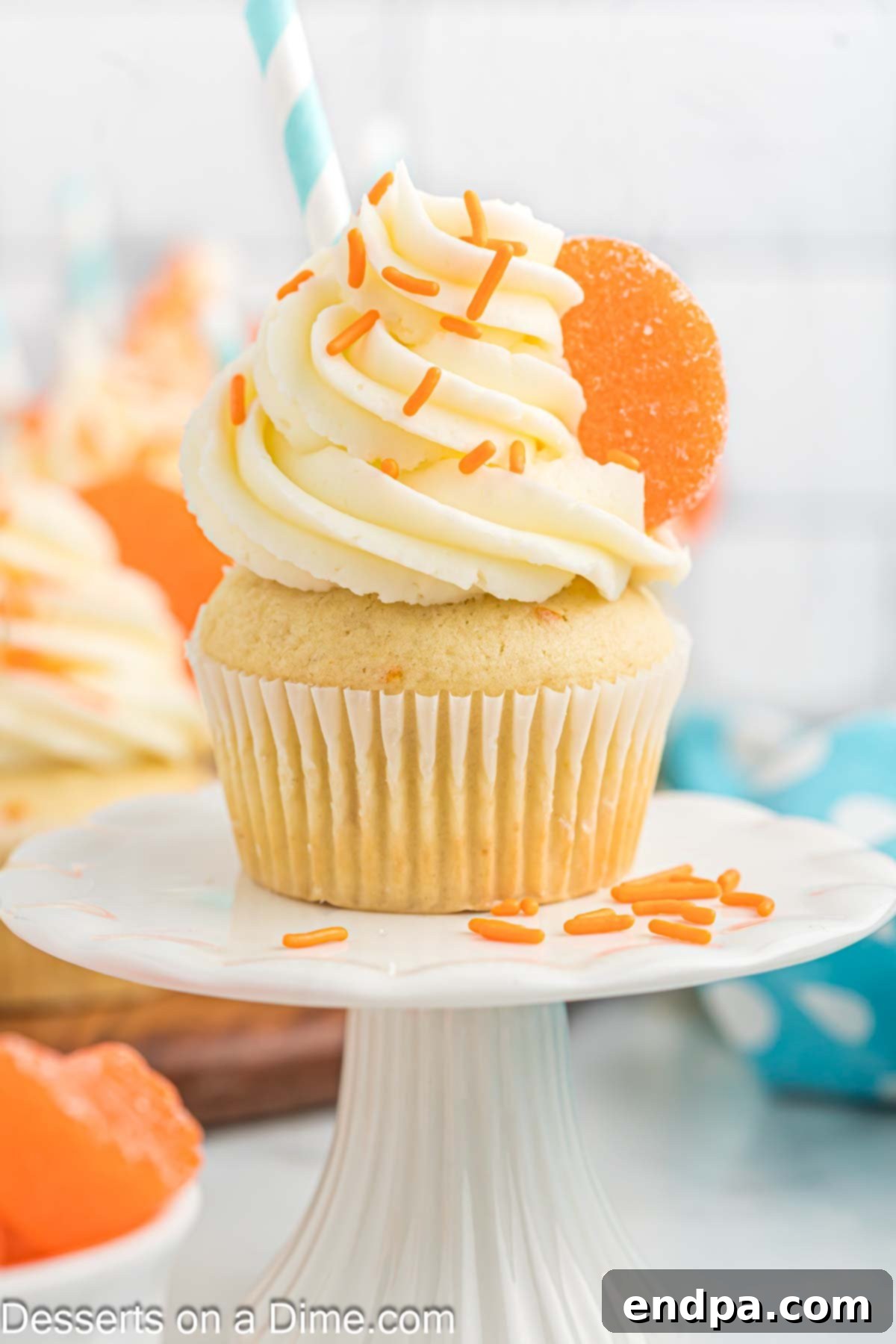 A beautifully frosted orange creamsicle cupcake on a white cake stand, garnished with an orange slice and sprinkles.